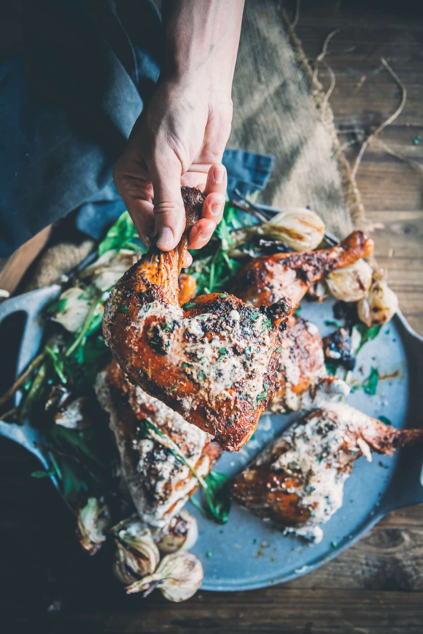 Hand holding smoked chicken leg quarter thats been slathered in Alabama white sauce over a platter of more chicken. 