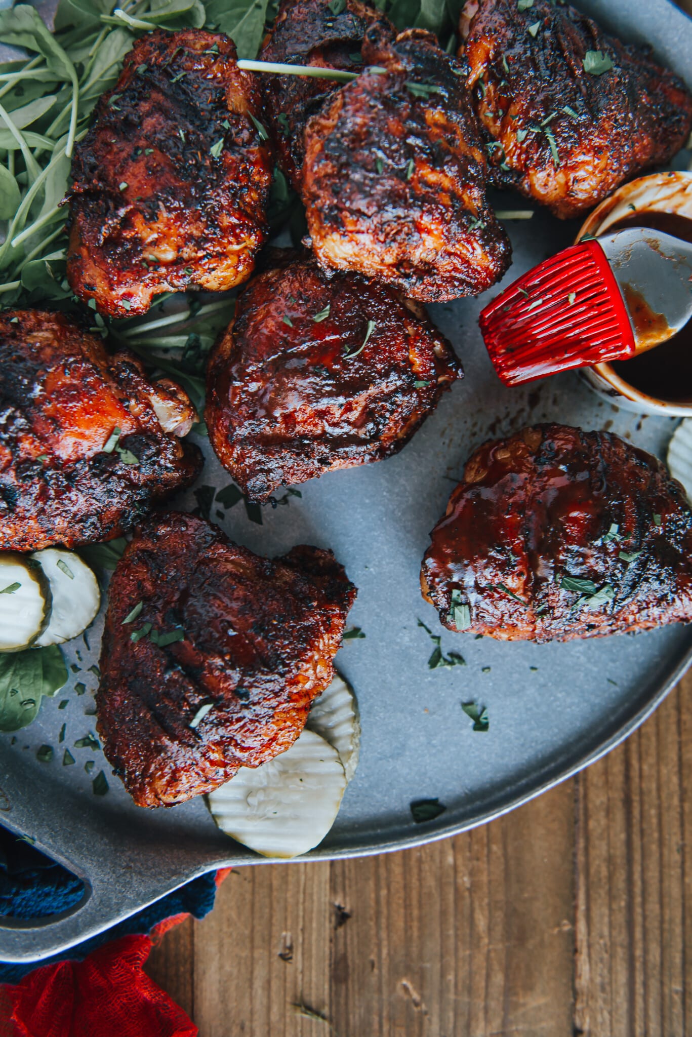 BBQ chicken thighs on a platter with pickles and barbecue sauce. 