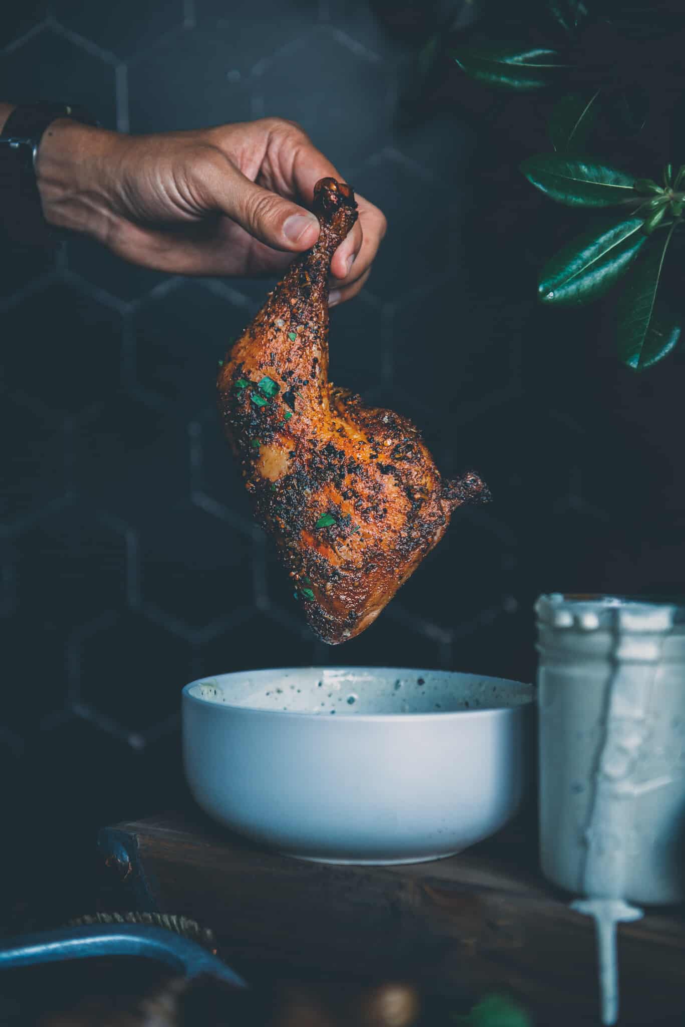 Hand holding smoked chicken leg quarter over a bowl of sauce. 