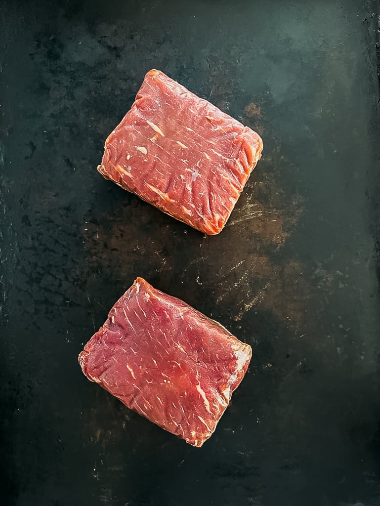 2 petit top sirloin steaks, raw, on a black baord.