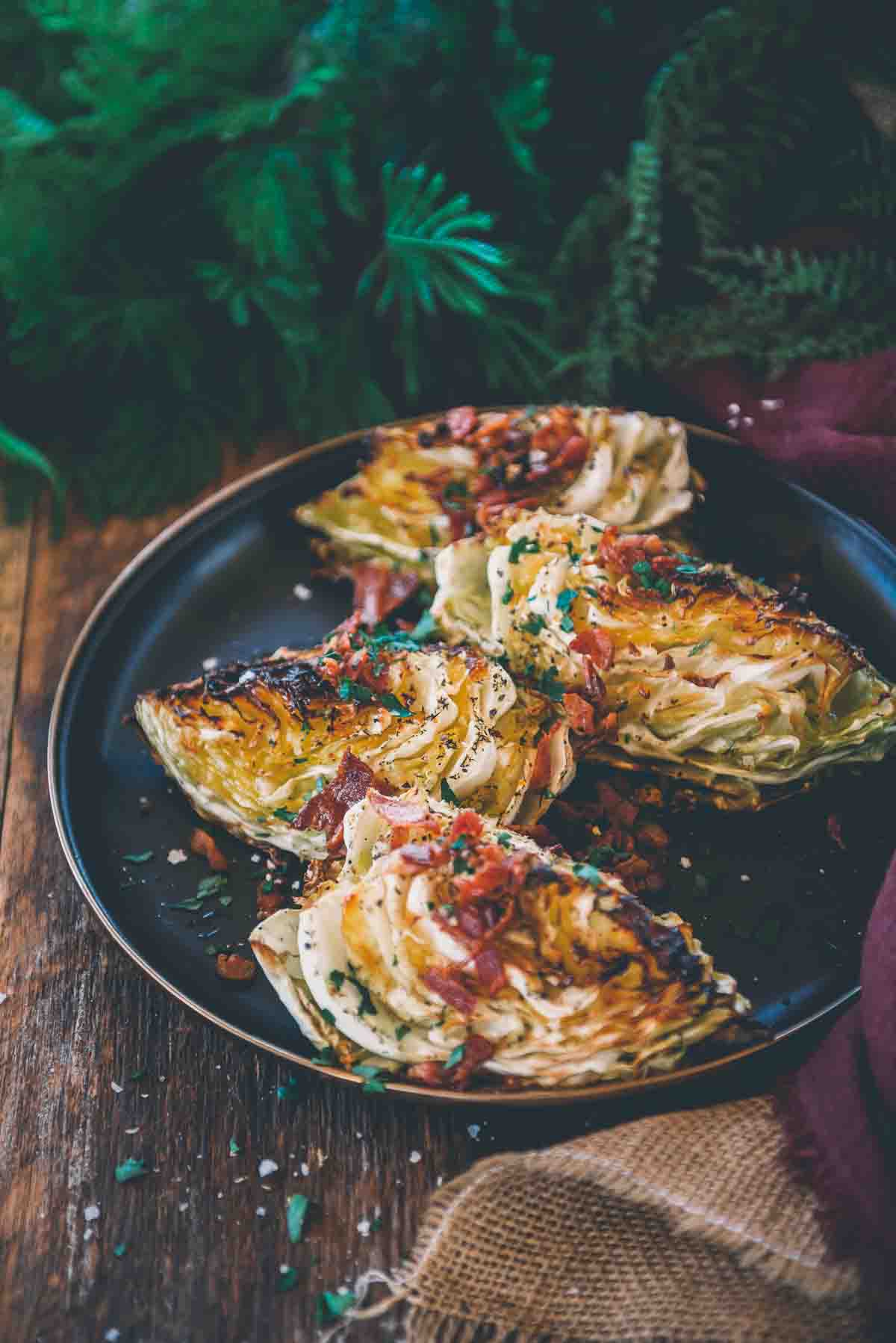 Black platter with 4 wedges of smoked cabbage with crispy golden brown edges garnished with crumbled bacon. 