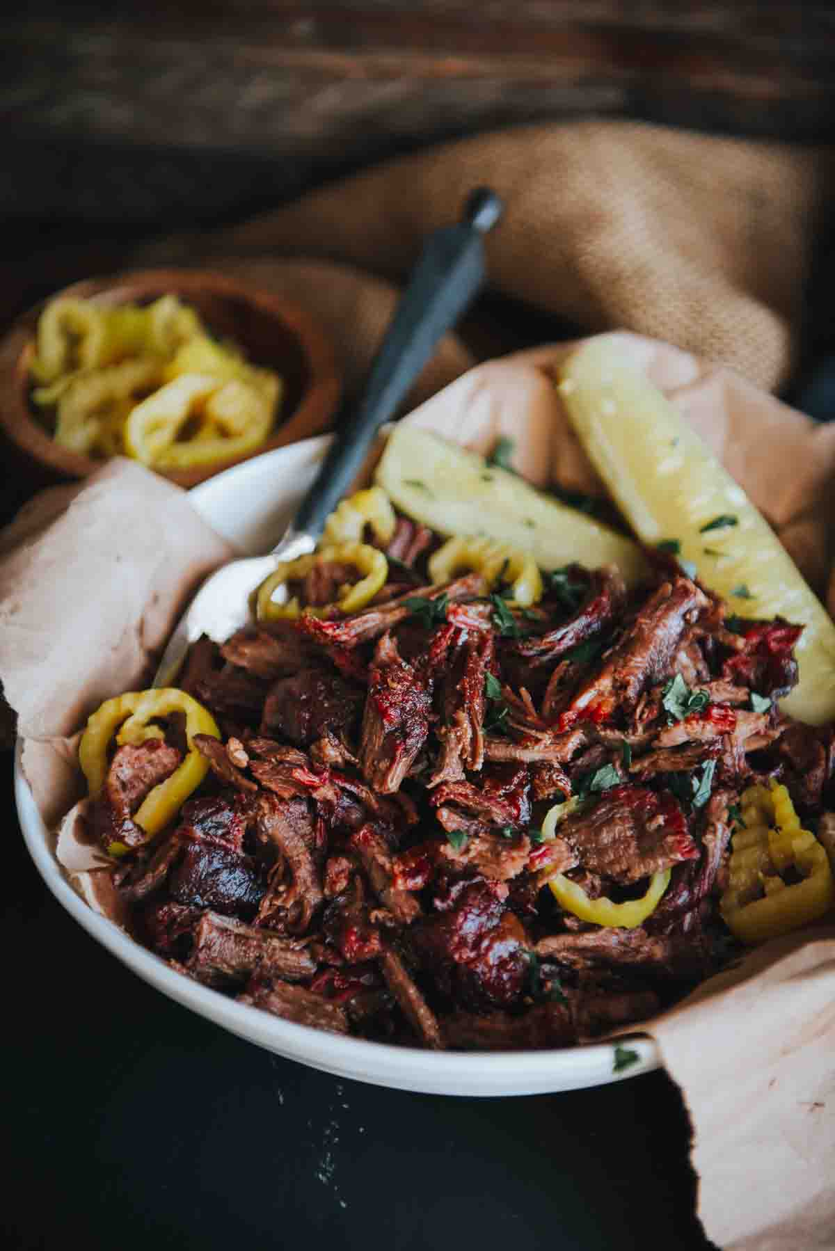 A bowl of pulled beef, showing perfect red color from being smoked, with a fork for serving and pepperoncini as garnish. 