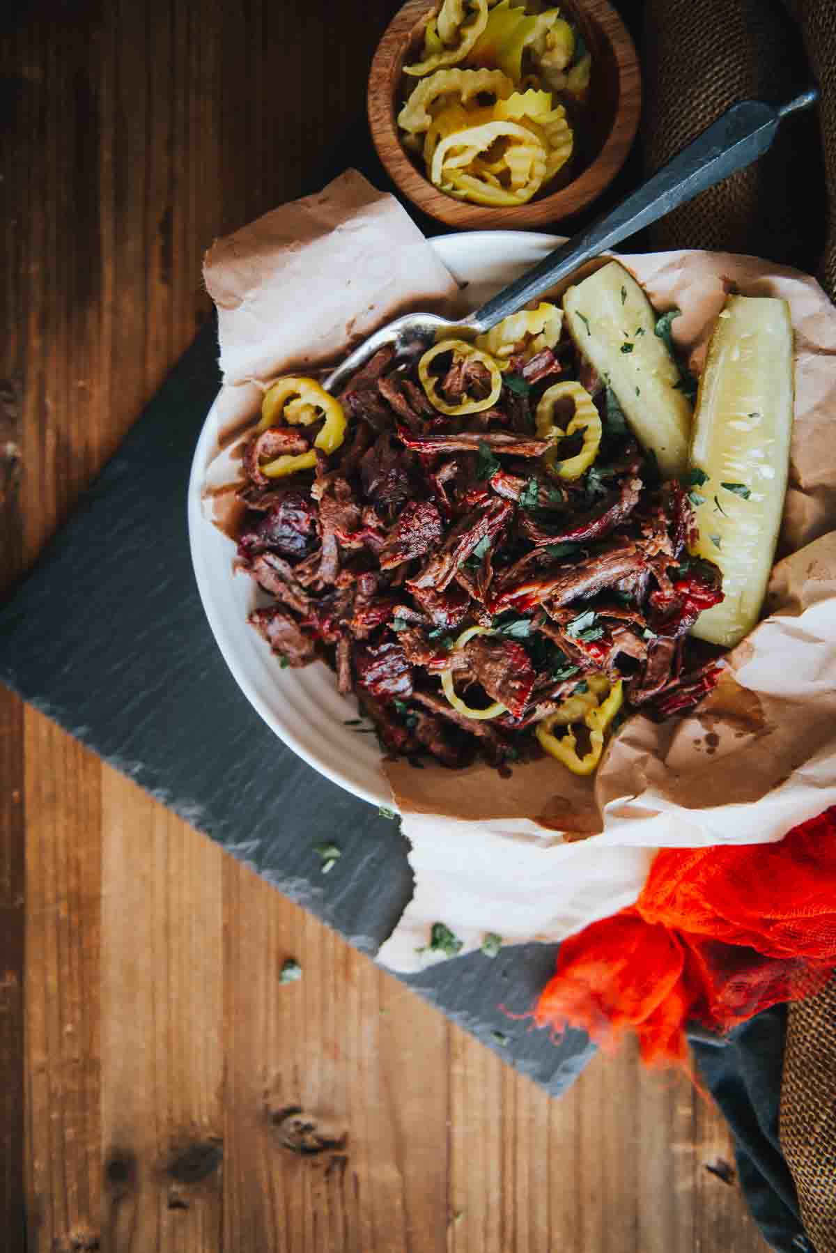 Overhead shot of smoked chuck roast thats been shredded to show a pink smoke ring on pulled beef in a bowl with pepperoncini peppers and pickles. 