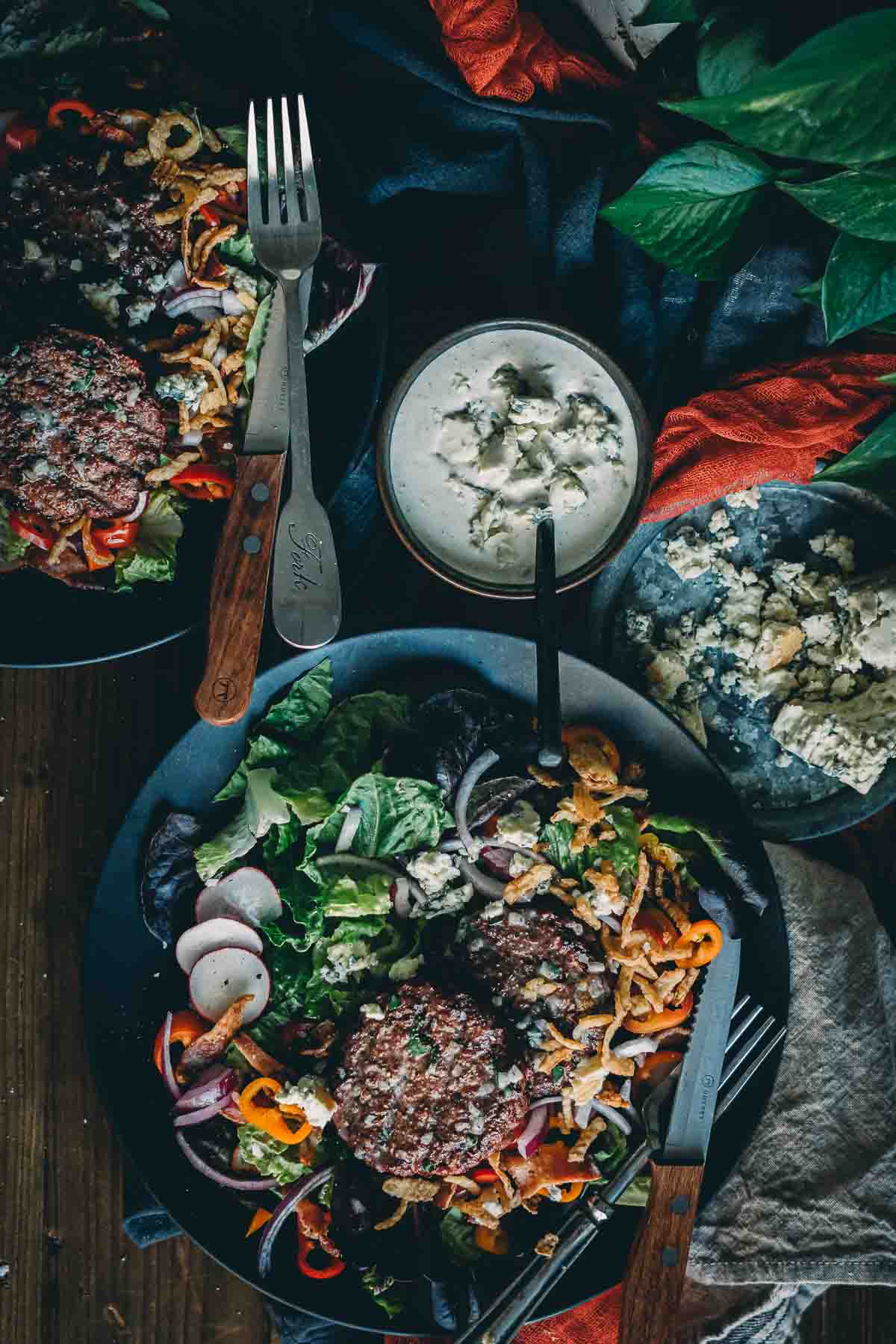 Above shot of 2 plates filled with smoked burger patties over tossed greens and veggies with blue cheese dressing in jar on the side and crumbles. 