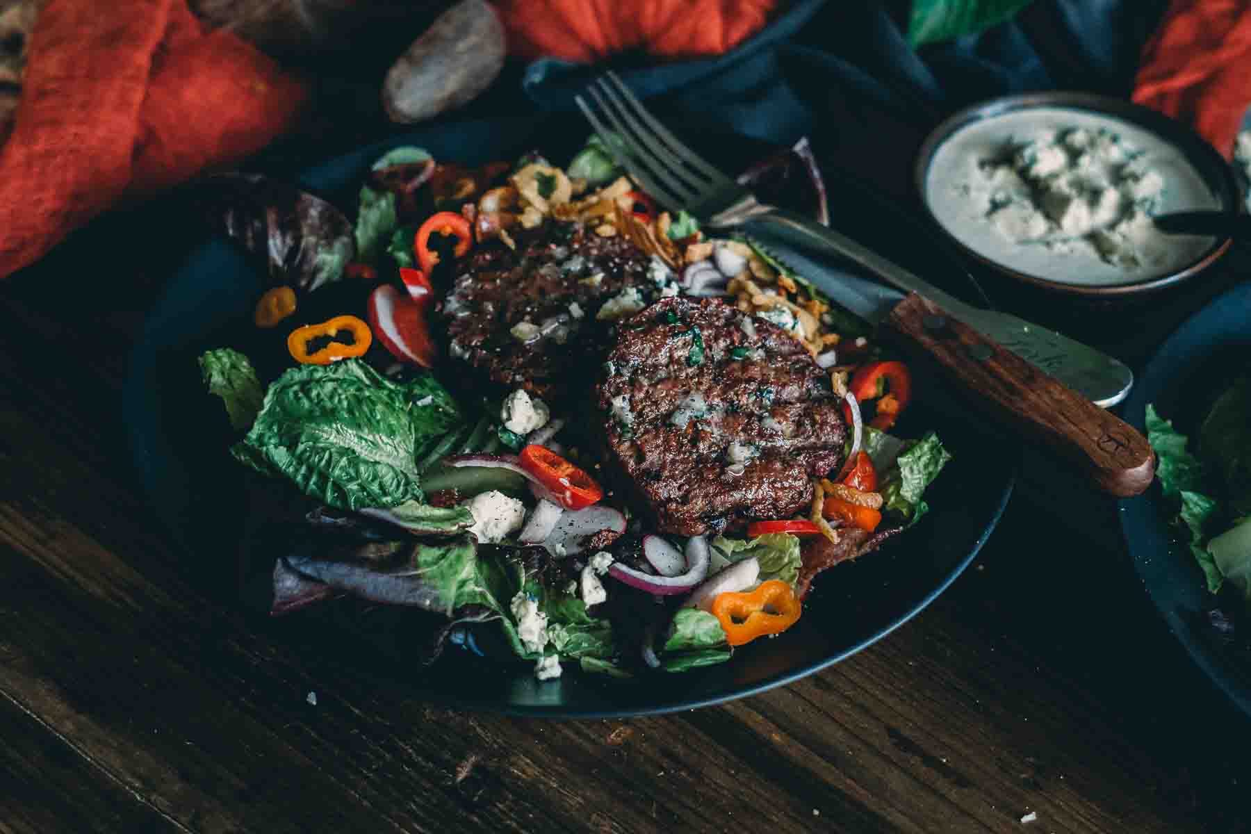 Side shot of steakhouse smoked hamburger salad with bowl of blue cheese dressing with sliced veggies. 