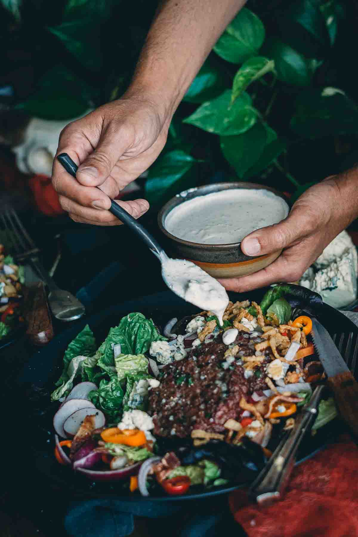 Hand drizzling homemade blue cheese dressing over smoked hamburger salad. 