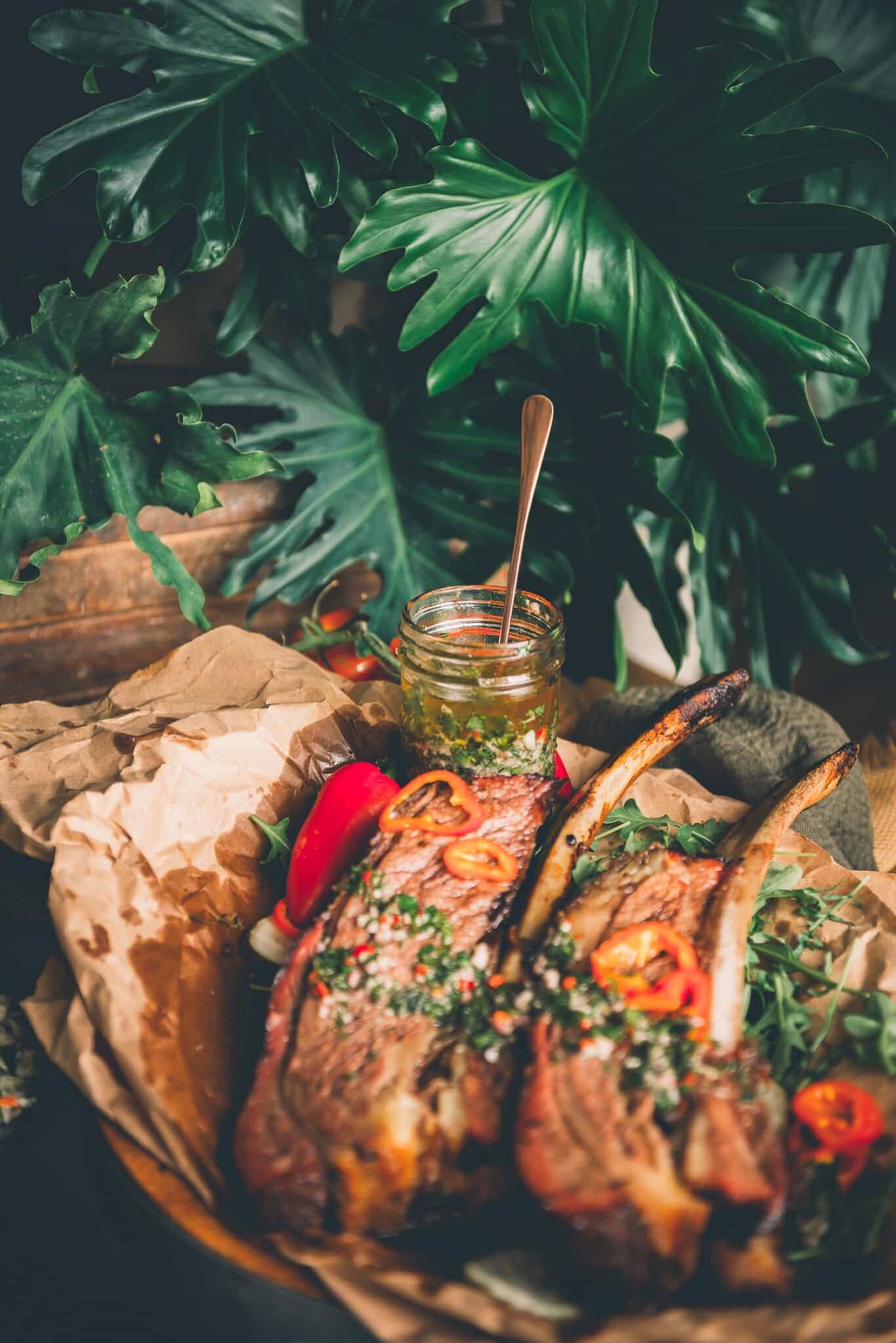 Jar of chimichurri nestled behind beef ribs in front of a luscious green plant. 