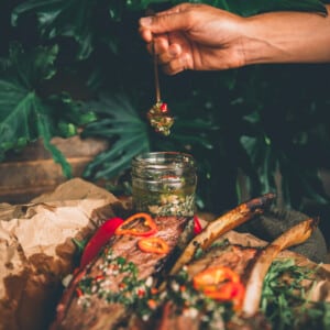 Hand holding spoon full of chimichurri over a jar with beef ribs slathered in the sauce in the foreground.