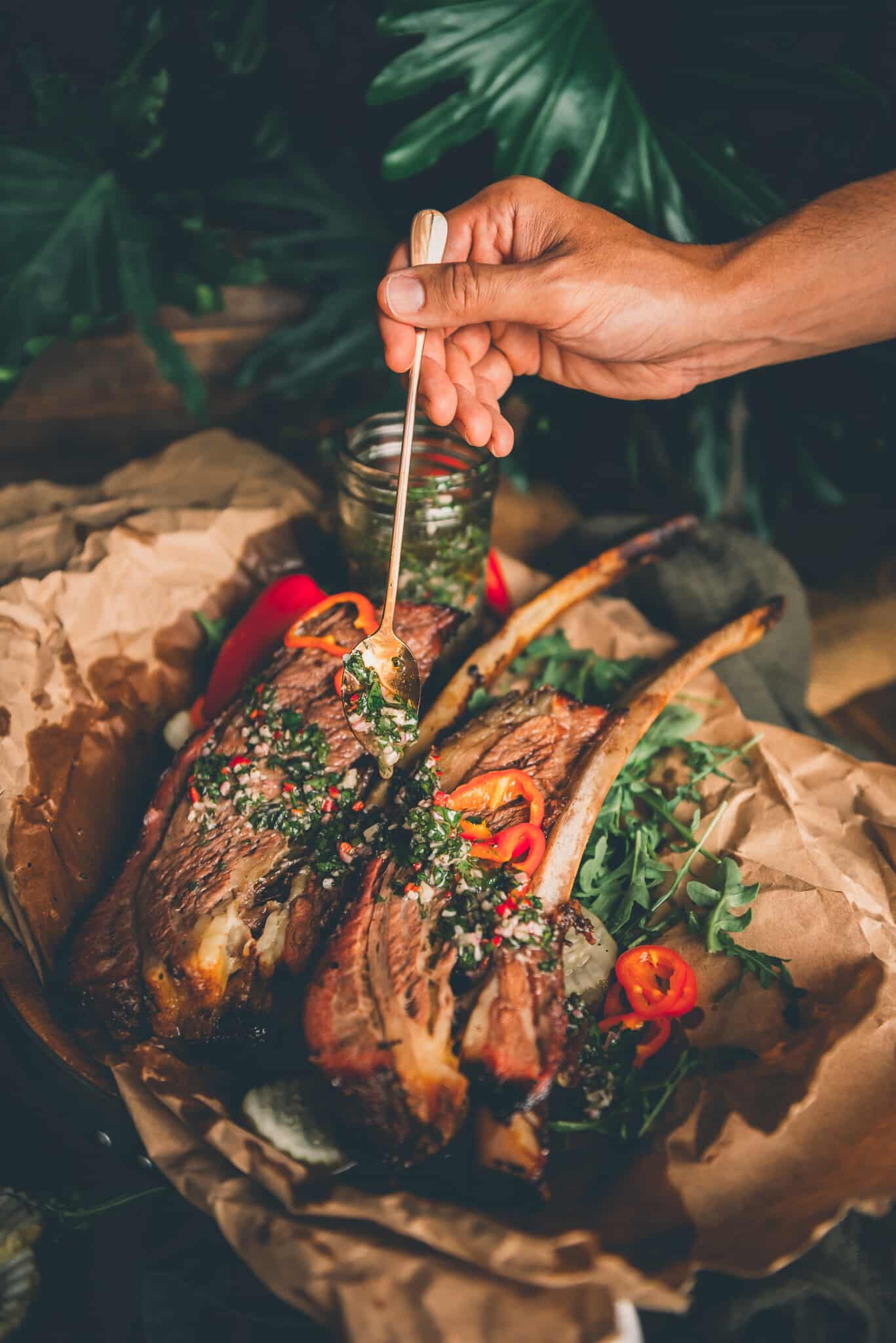 Hand drizzling chimichurri over big cuts of beef on crumpled paper. 