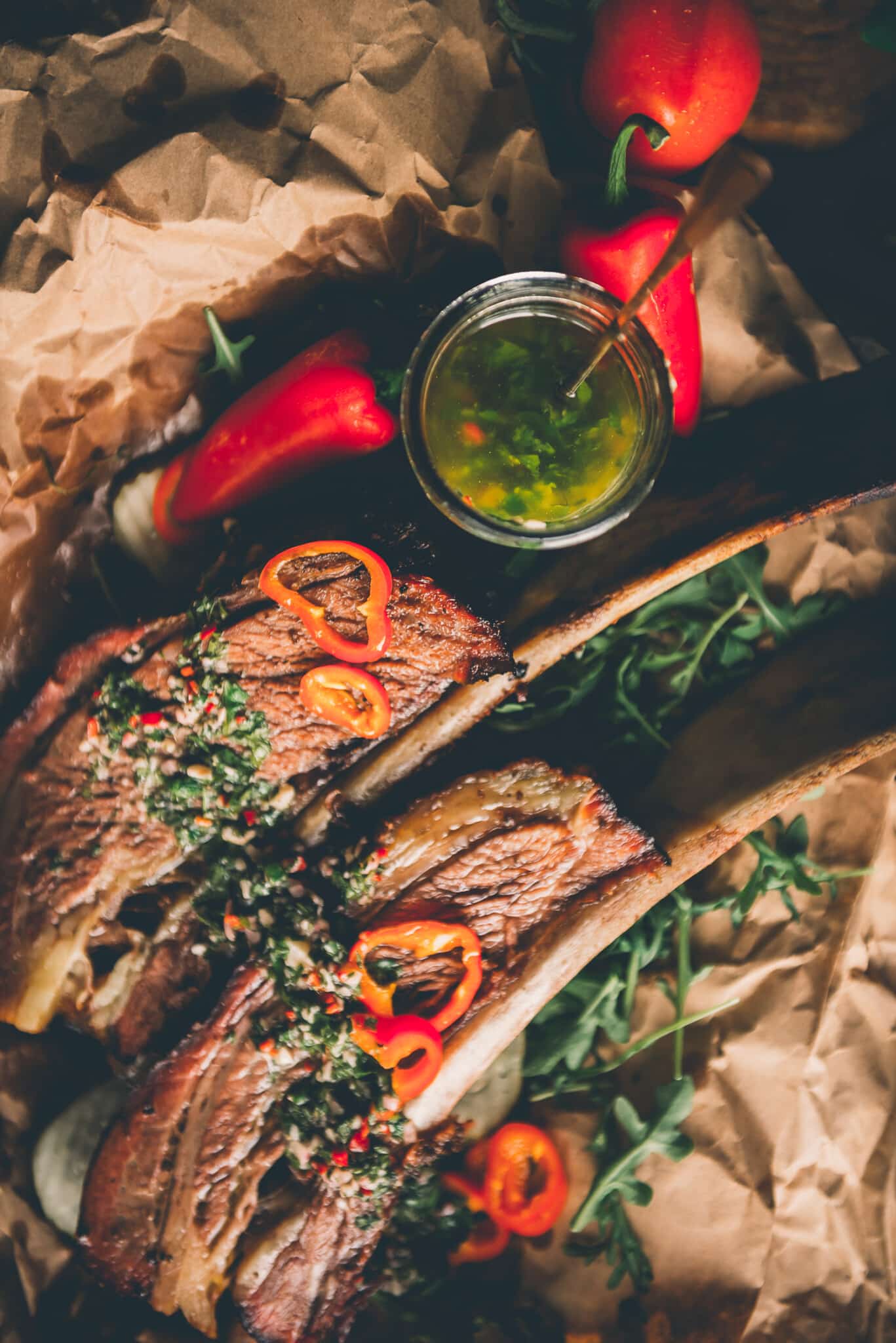 Above shot of homemade chimichurri in a mason jar next two two huge beef ribs slathered with the herb sauce and garnished with sliced peppers. 