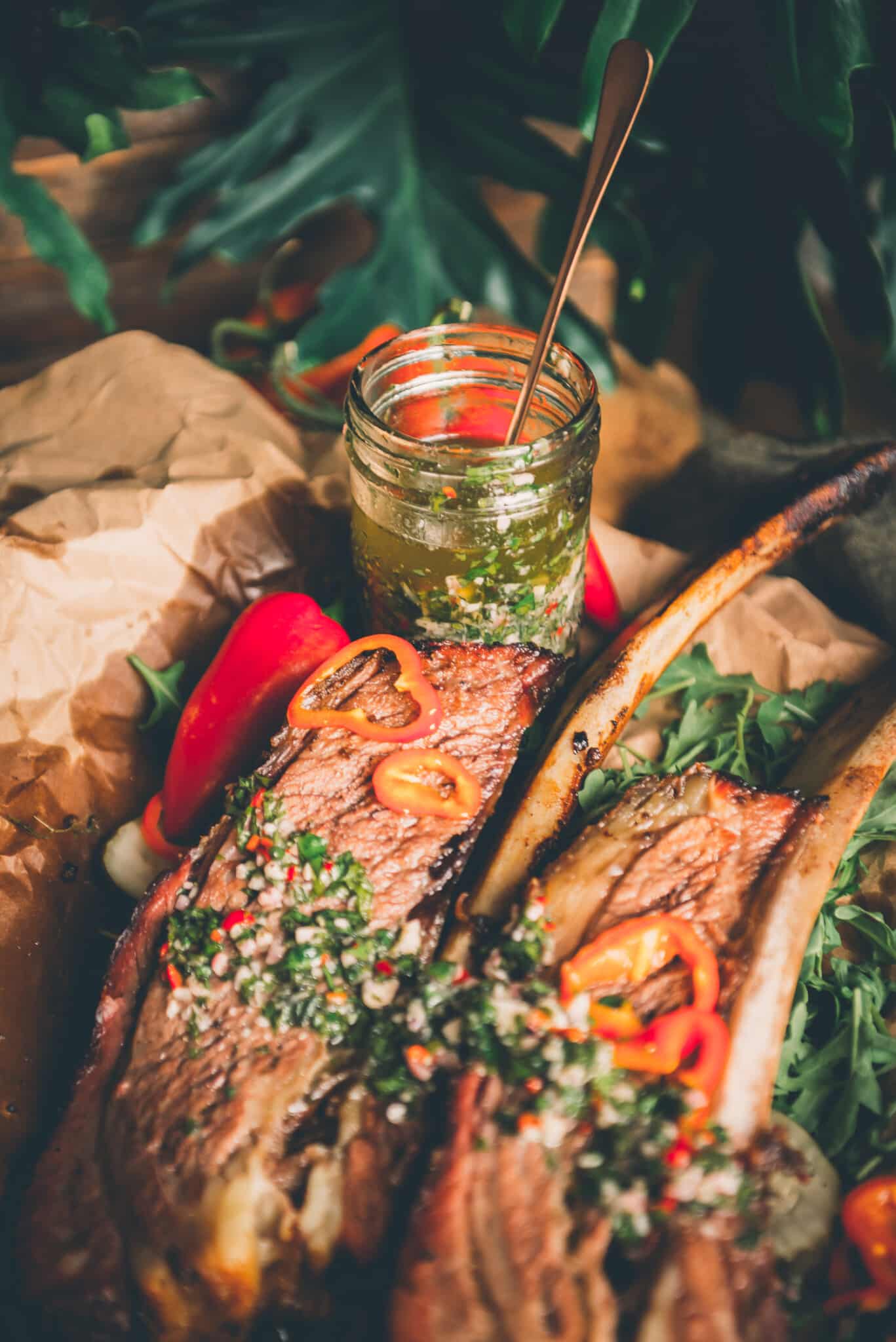 Jar of homemade fresh herb filled  with chimichurri with two beef ribs slathered in the sauce in foreground. 