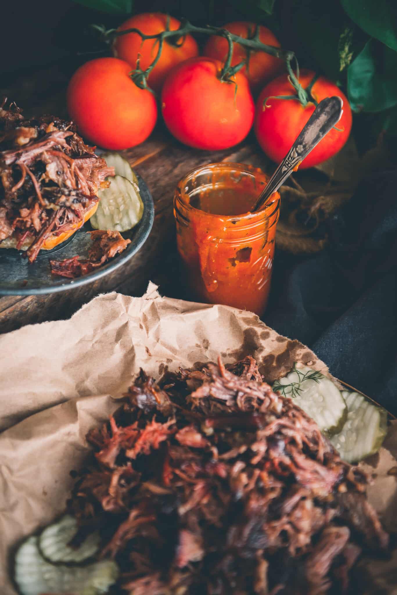 Messy jar of barbecue sauce with a spoon handle exposed nestled into a full table filled with shredded beef and bbq sandwich. 