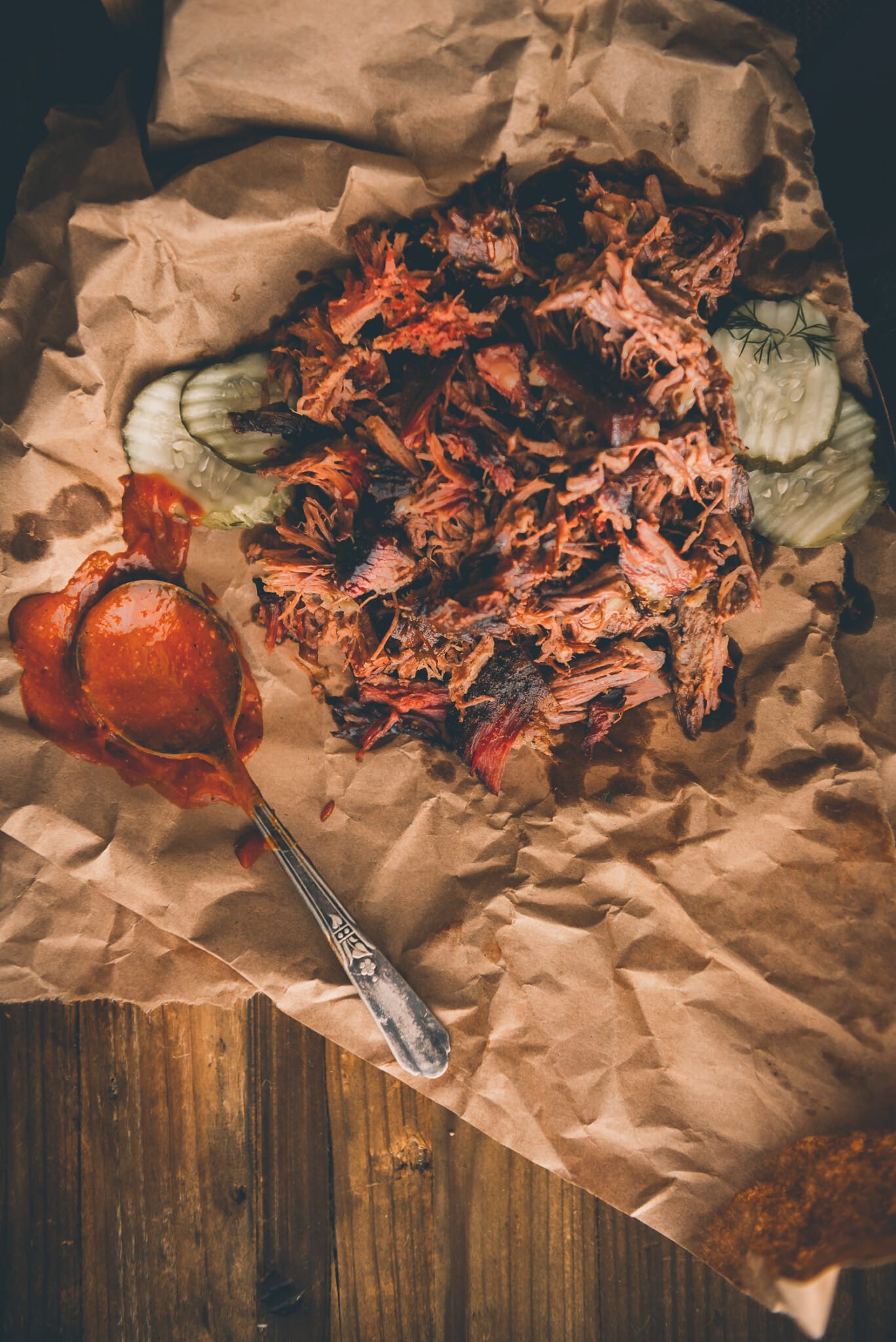 Shedded barbecued beef on a crumpled brown paper with pickles and spoon dripping with bbq sauce on the side. 