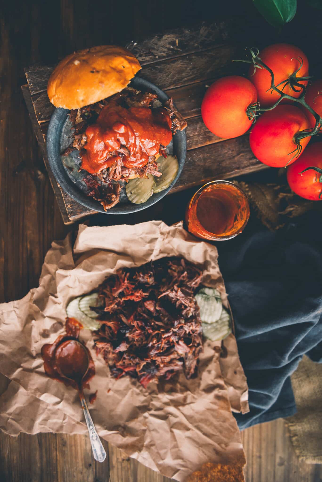 Overhead shot of pulled beef on a platter and on a small plate on a bun with pickles drenched in BBQ sauce. 