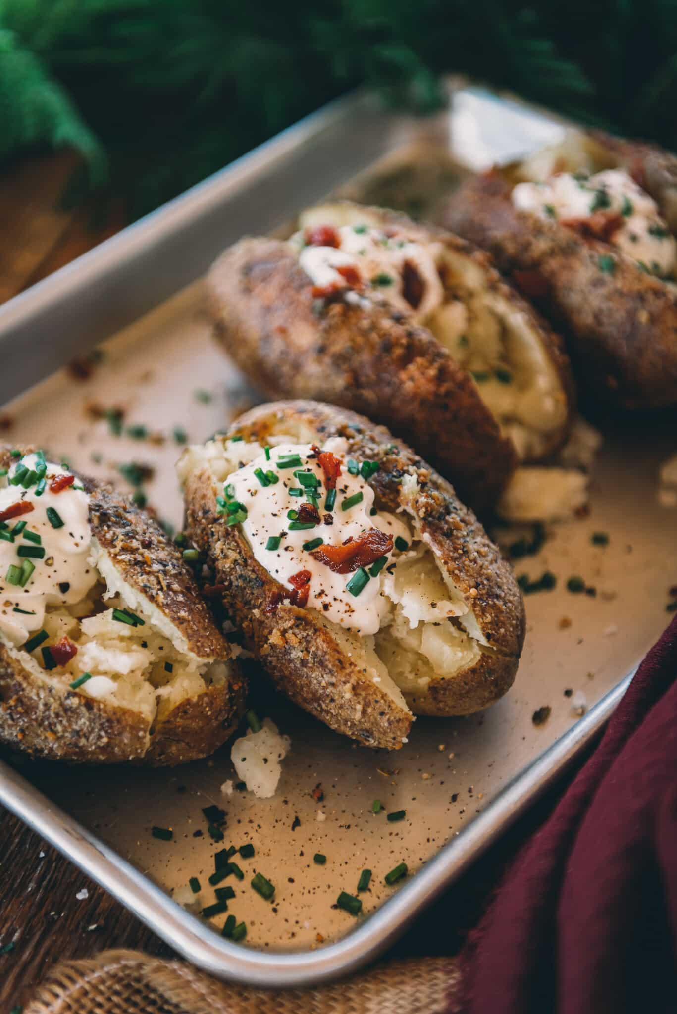 Close up of smoked baked potatoes showing herb crusty crispy skin, sliced and topped with sour cream, chives, pepper and bacon crumbles. 