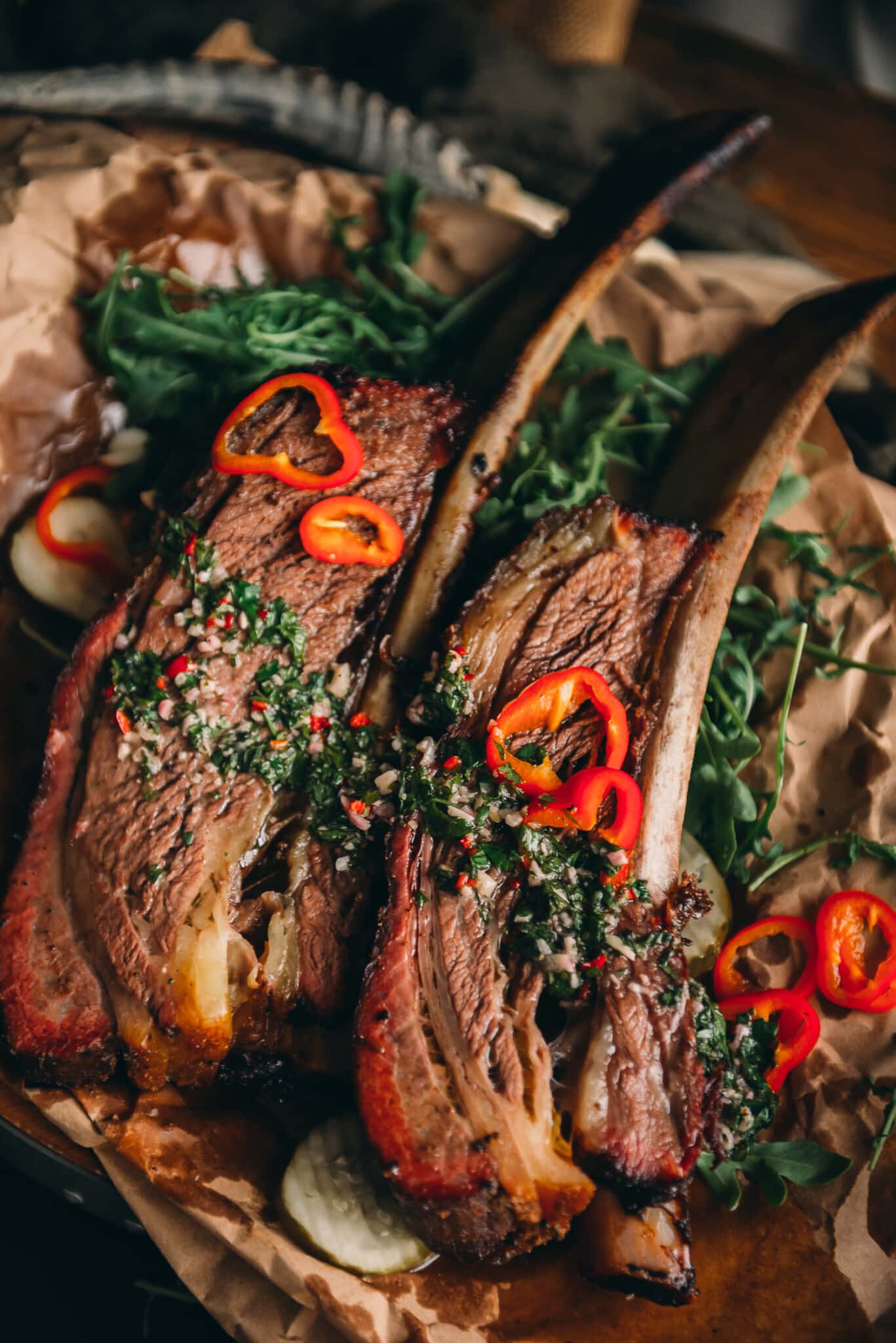 2 bone in beef ribs on a platter, garnished with bright green herb sauce and red peppers. 