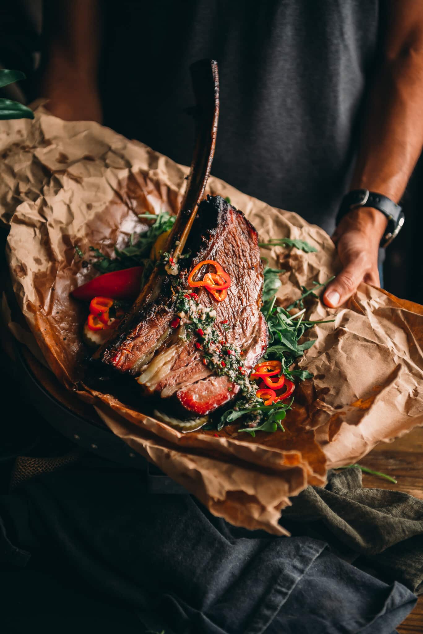 Hand holding juicy beef rib on a platter garnished with pepper slices, arugula, and chimichurri. 