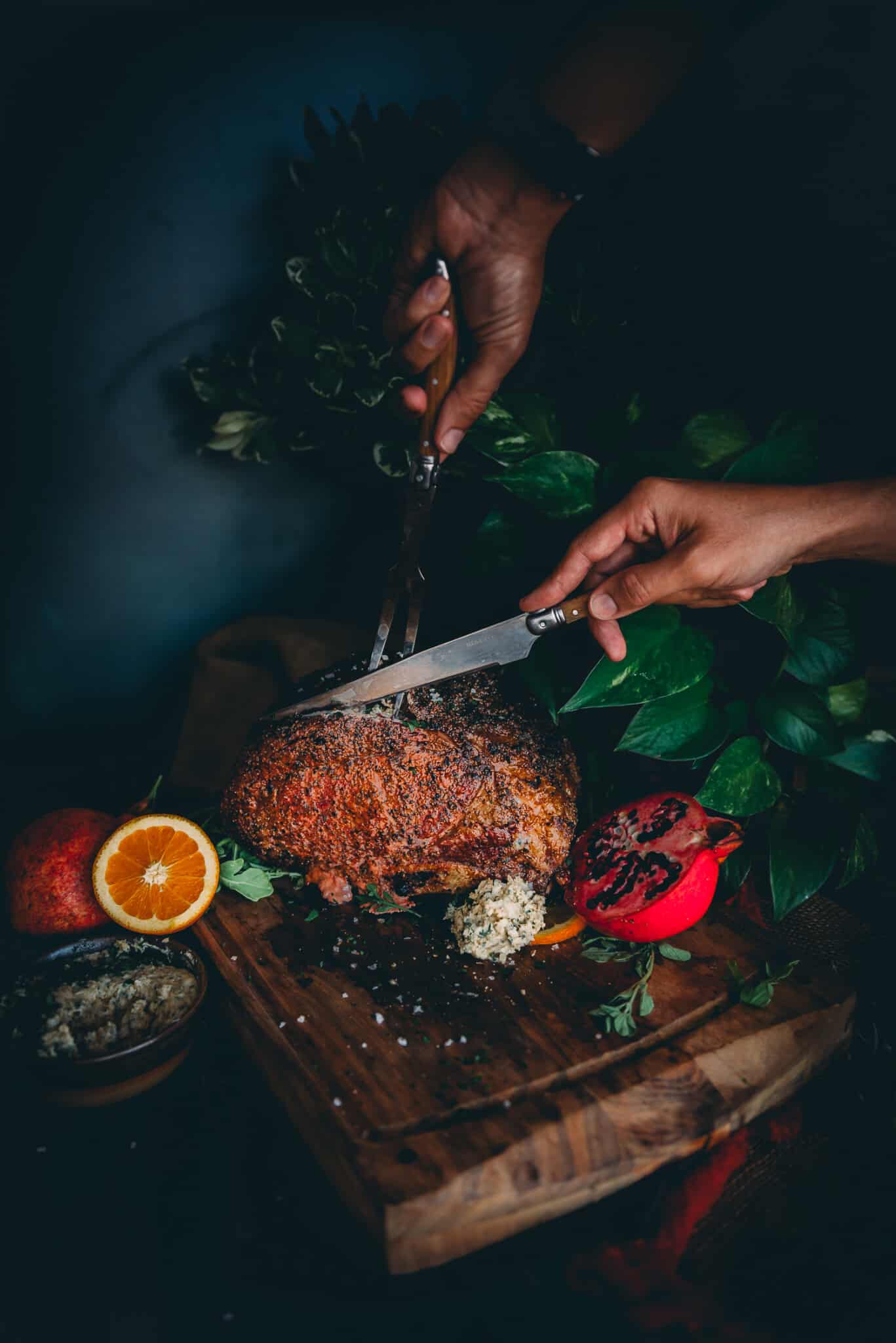 Hands holding carving fork and knife cutting into a cooked prime rib. 