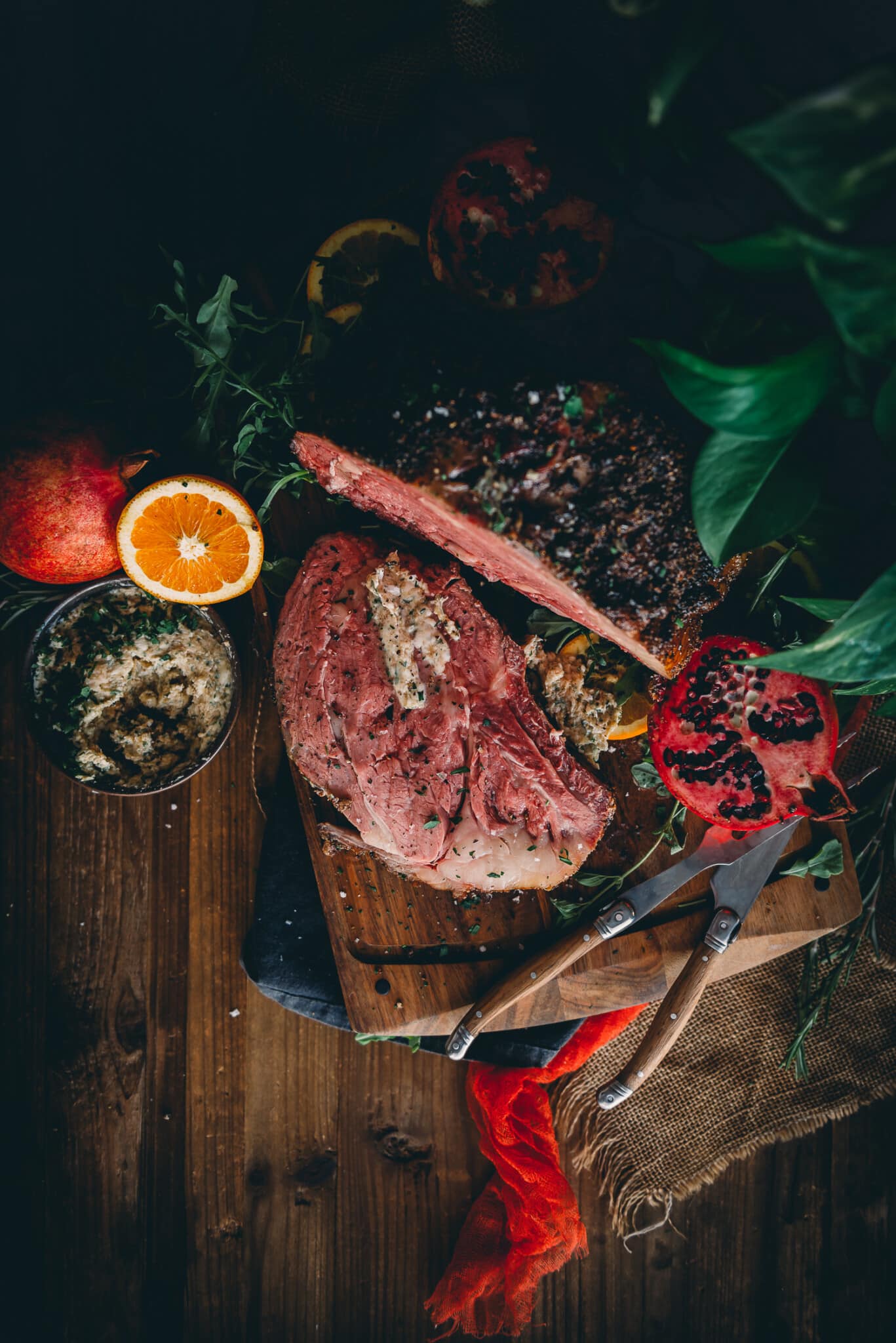 Colorful overhead shot of smoked prime rib, with one slice, steakhouse herb butter, utensils and seasonal citrus and pomegranate. 