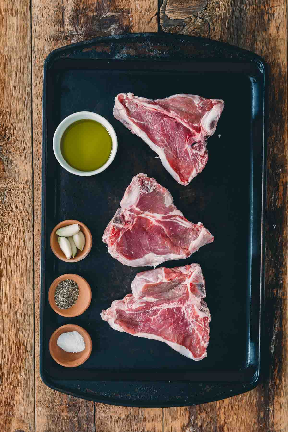 Raw veal chops arranged on a black baking sheet with salt, pepper, and garlic. 
