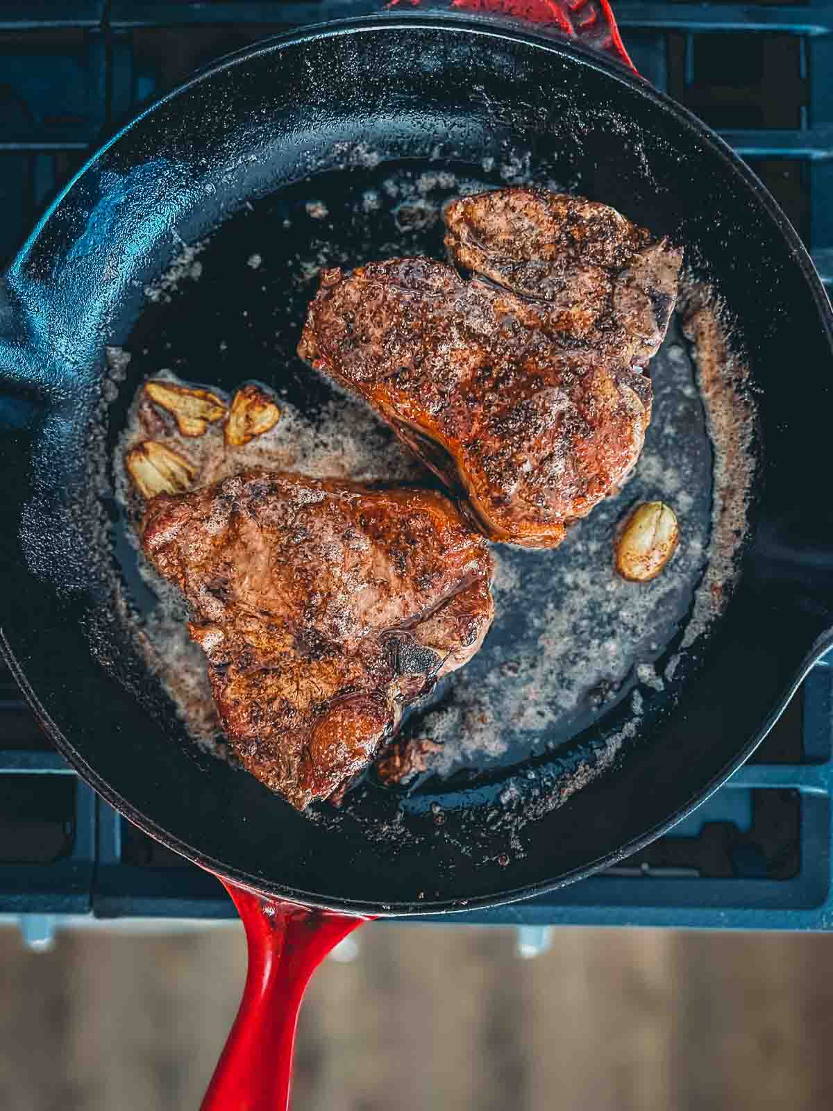 Two veal chops in a cast iron skillet with garlic, bubbling oil and butter being pan seared. 