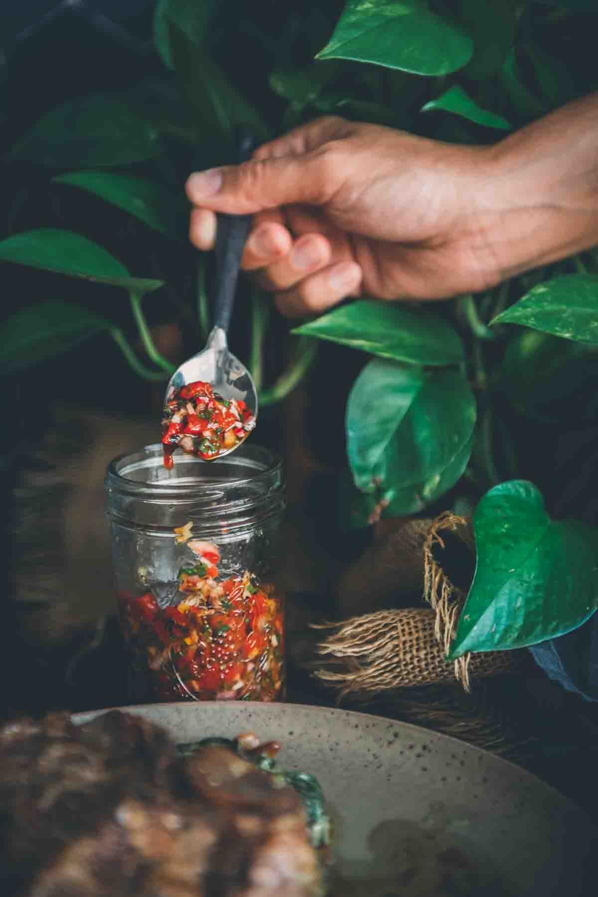 Hand pulling spoon out of jar filled with red chimichurri sauce. 