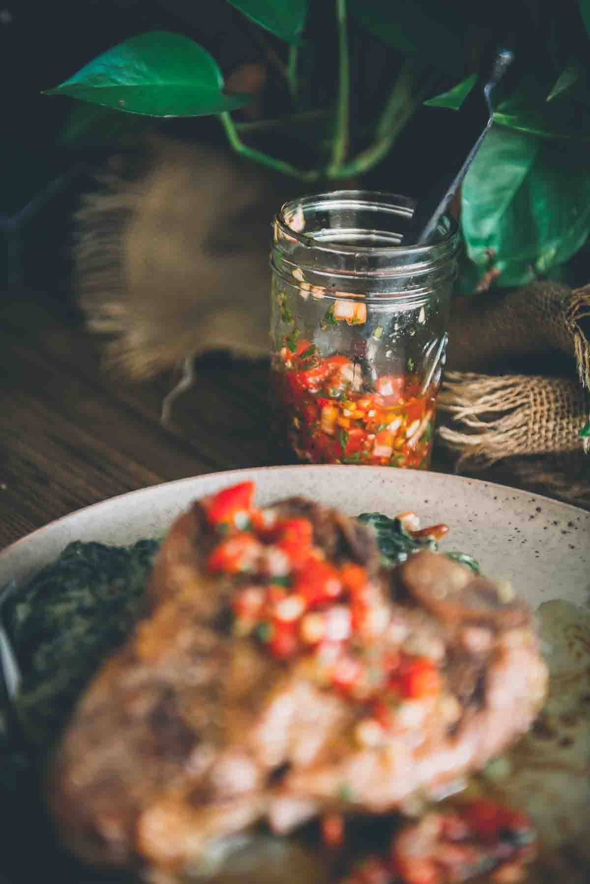 Jar of smoky red chimichurri sauce in focus behind meat with it spooned on top. 