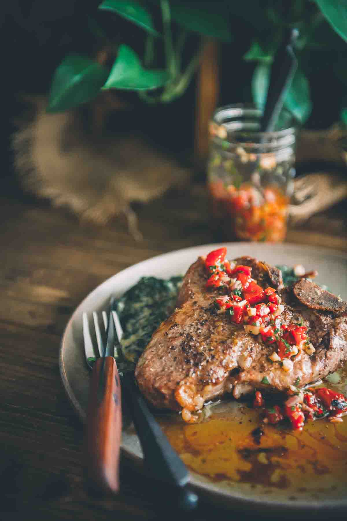 Cut of meat with red chimichurri on top in focus with jar of it in the background. 