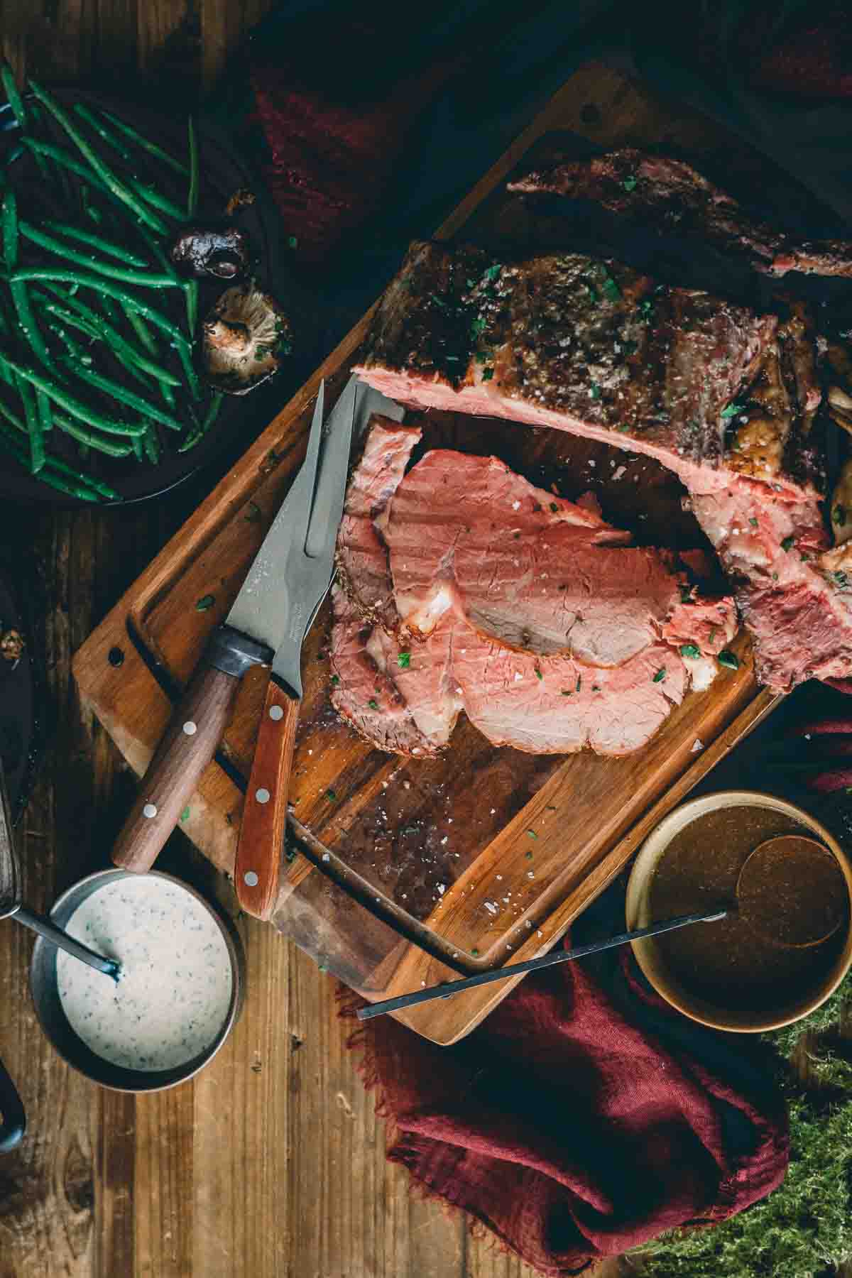 Overhead shot of prime rib sliced on a cutting board with jus and sauce in bowls near by. 