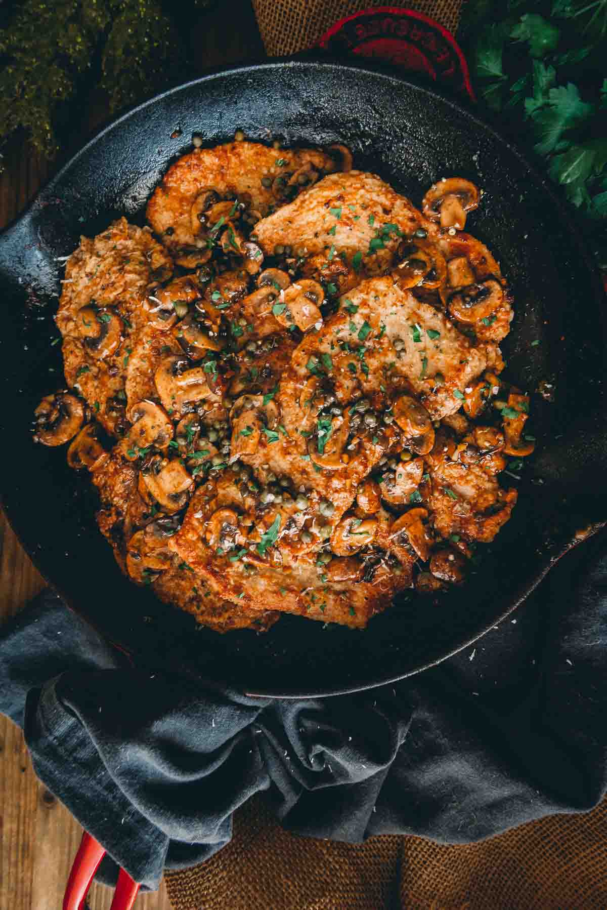 Overhead shot of skillet showing a pile of golden brown veal cutlets cooked with mushrooms and capers.