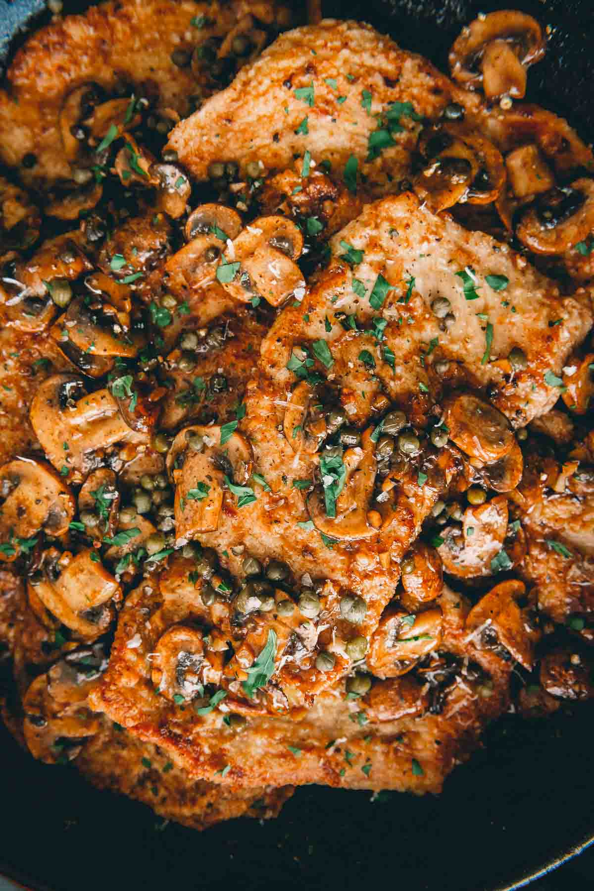 Close up of veal cutlets in a cast iron pan, coated in a marsala mushroom sauce with capers and freshly minced parsley. 