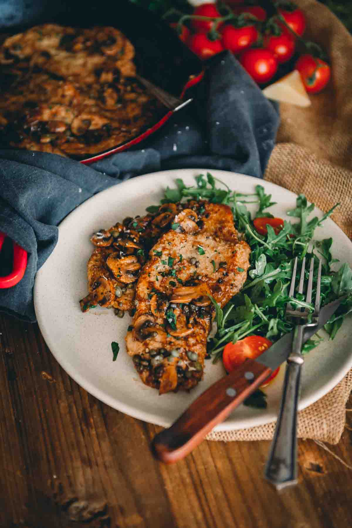 Plate with pan fried veal cutlets over arugula and sliced tomatoes with knife and fork. 