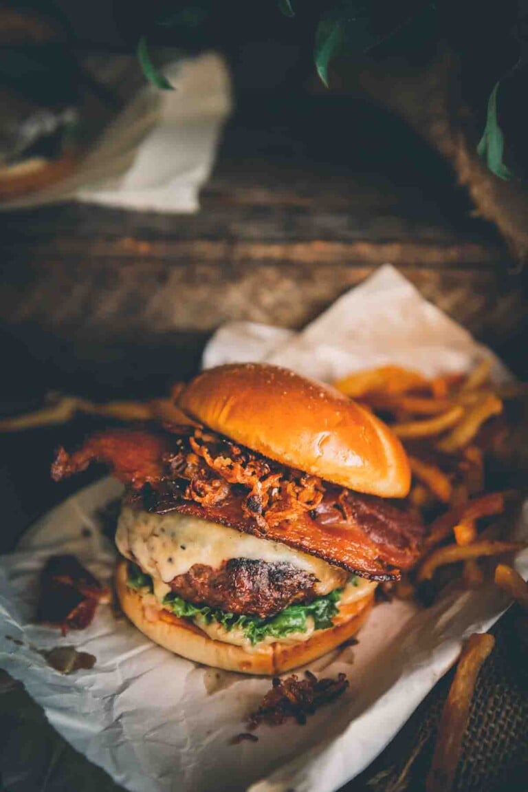 Huge ground venison burger on a soft bun with lettuce, cheese melting down the side, thick cut bacon, and crispy shallots with fries in the background.