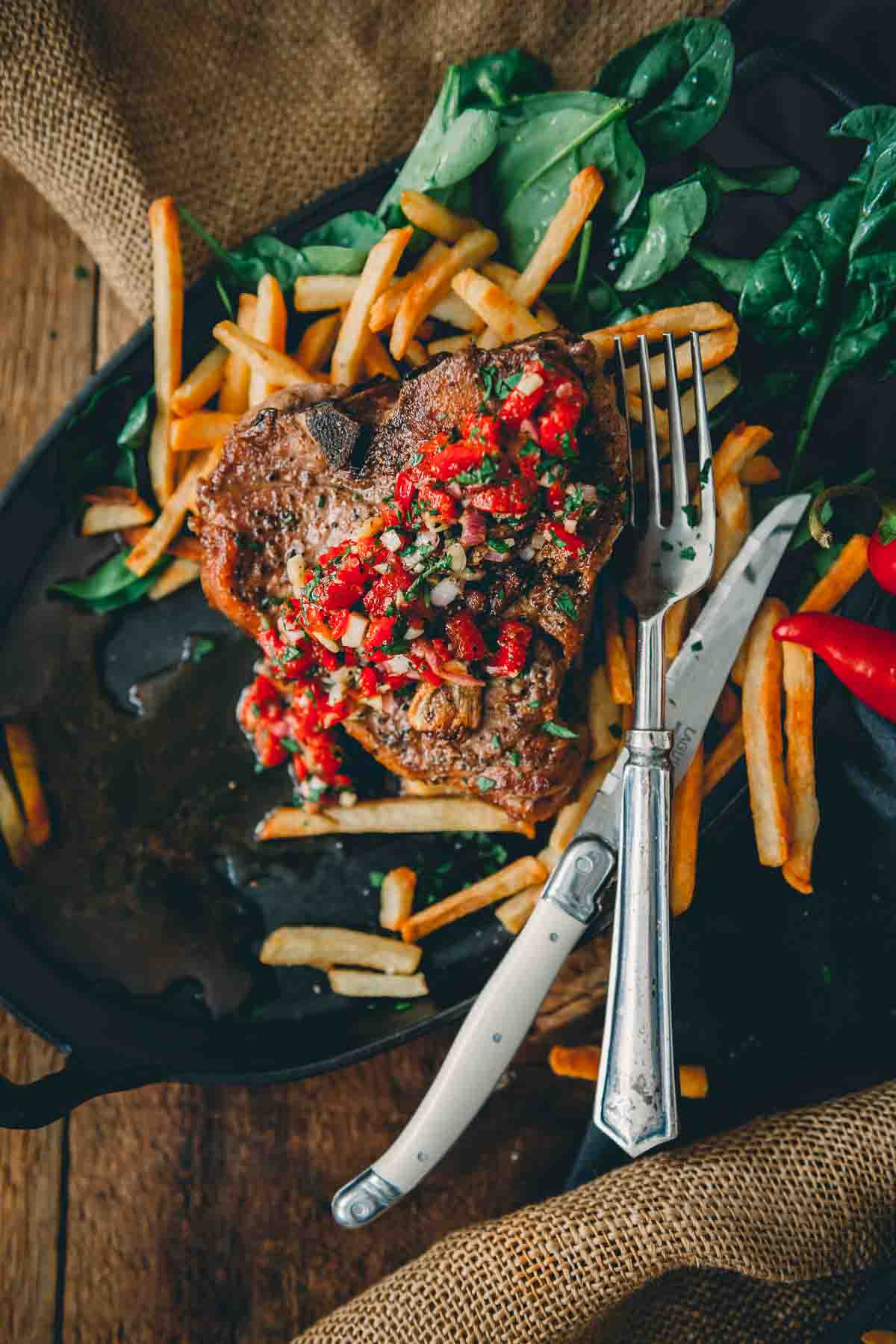 Vibrant photo of a pan seared veal chop with bright red chimichurri on top over golden fries and spinach. 