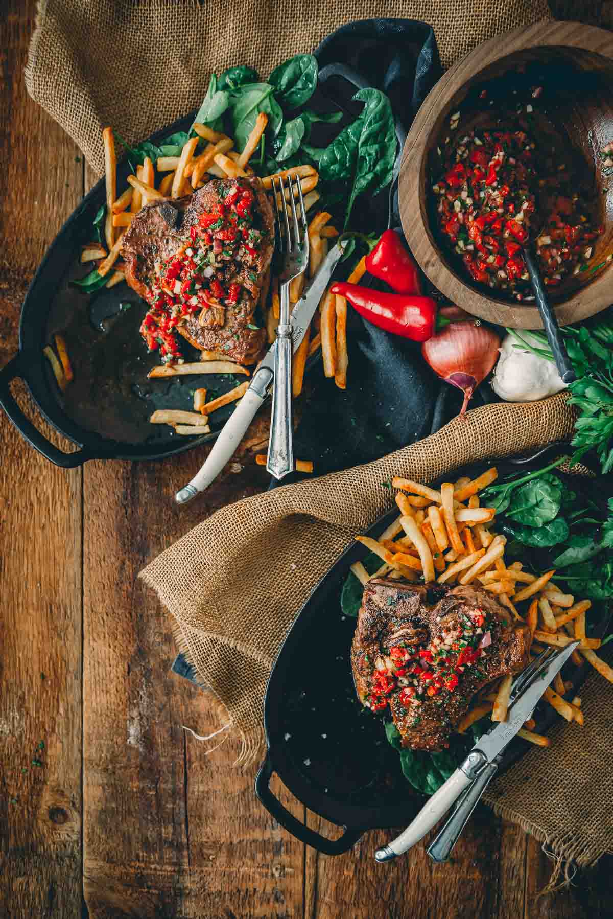 Overhead shot of two plates of beautifully seared veal chops with fries and salad and red chimichurri on top. 