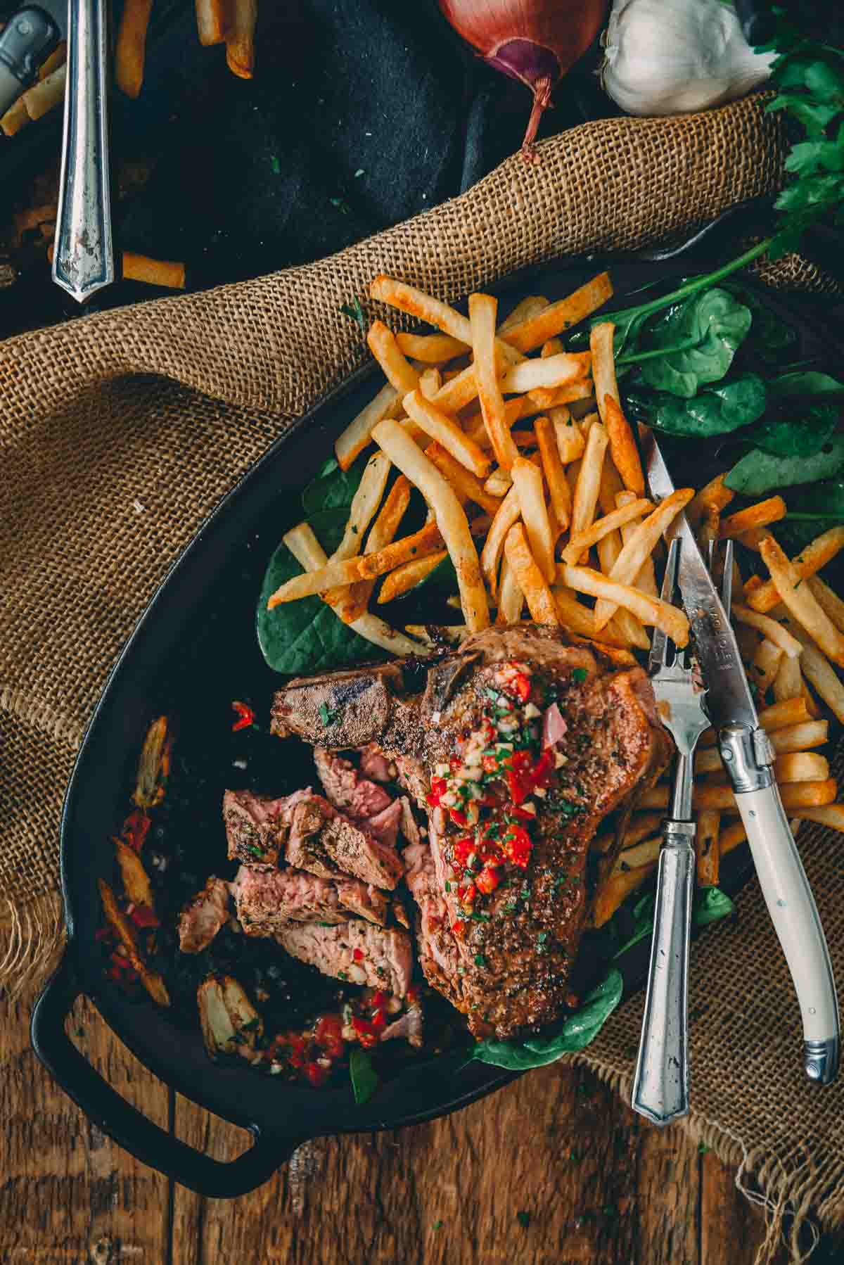 Veal loin chop with half sliced and other half covered in red chimichurri, nestled over fries and greens.