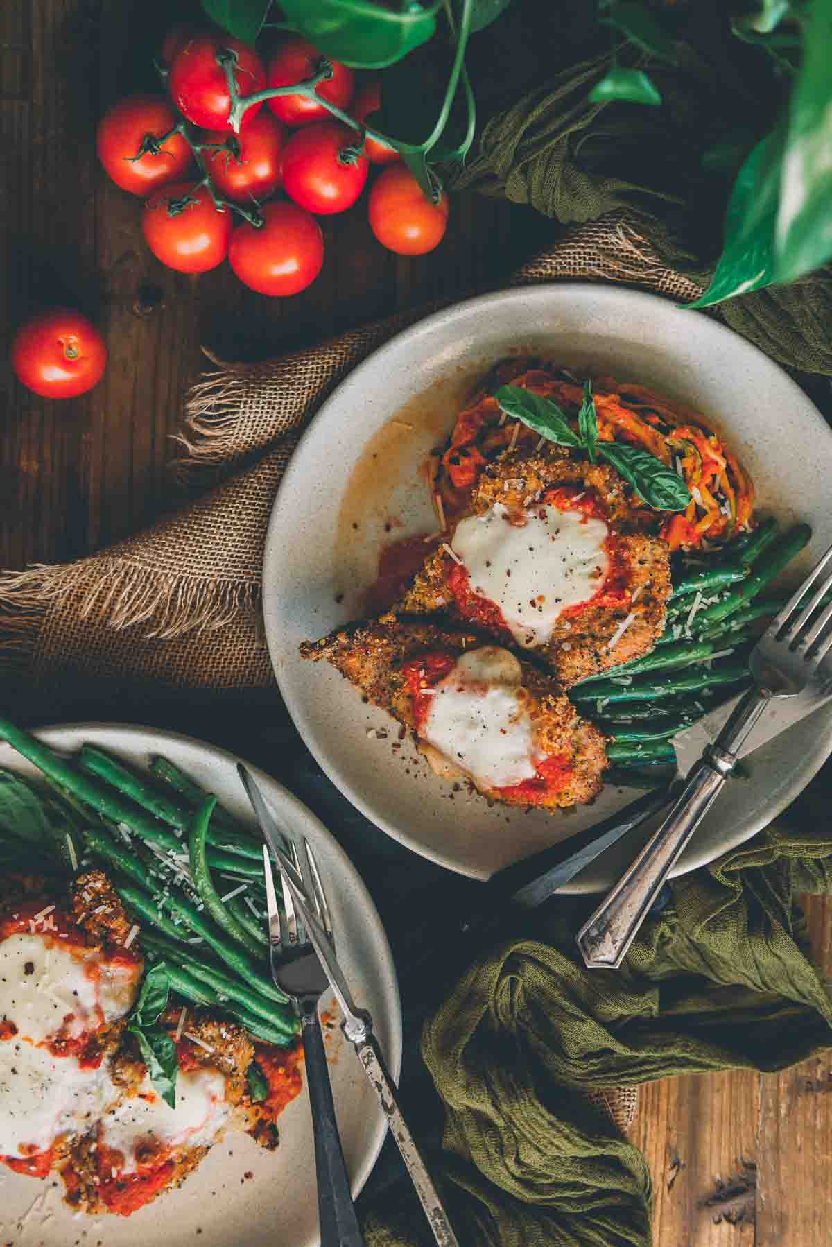 Overhead shot of veal parmesan on plates with green beans and zoodles in marinara sauce garnished with basil. 