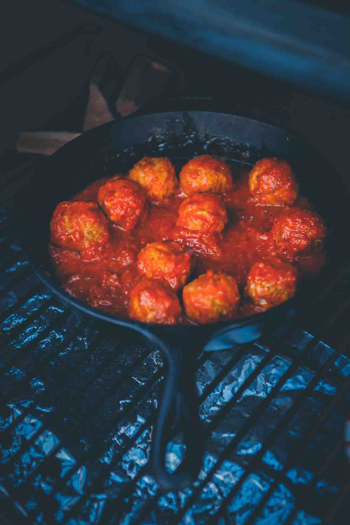 Cast iron skillet filled with meatballs coated in marinara sauce on grill grates. 