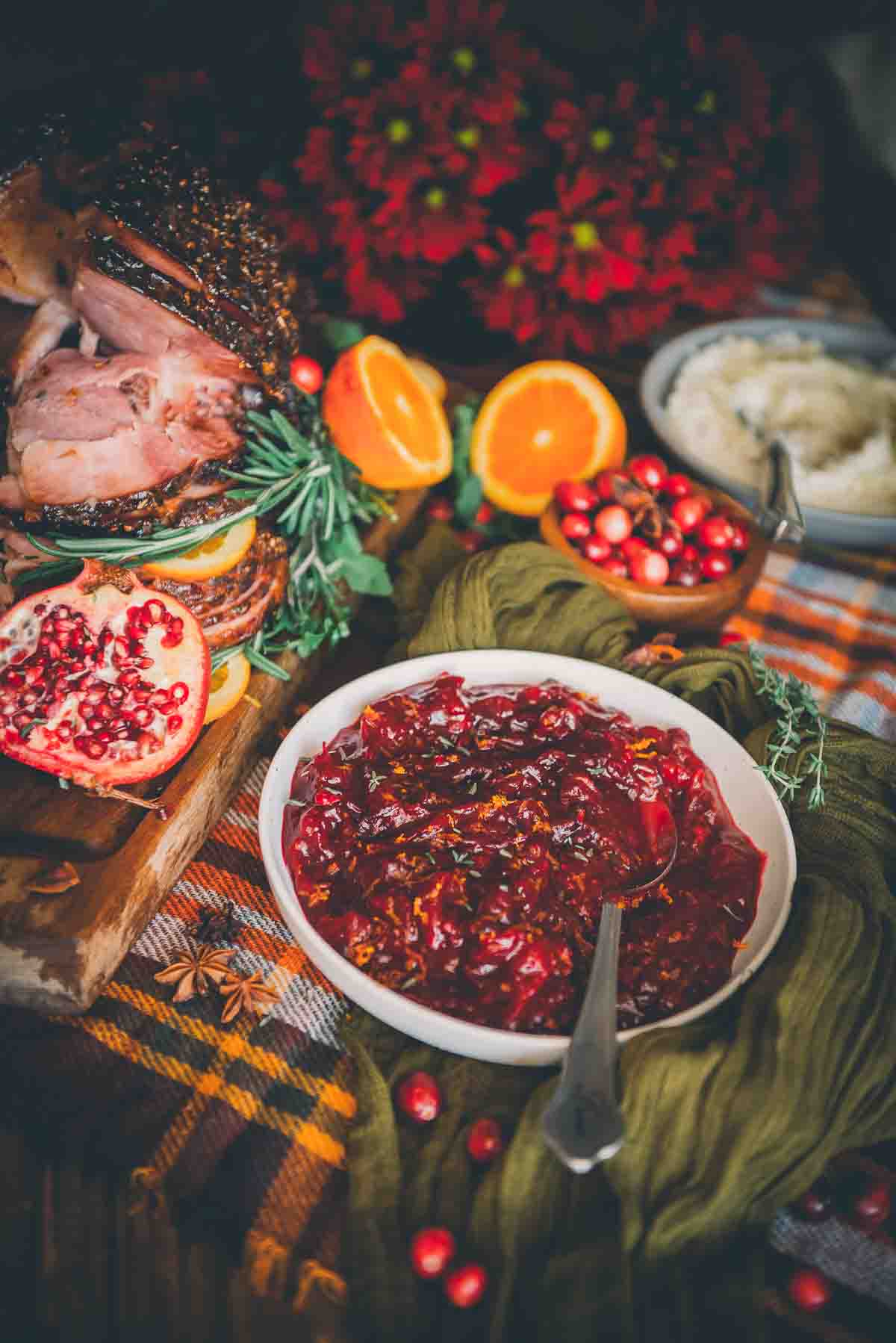 Festive colorful table with bowl of homemade cranberry sauce in a white bowl with orange zest and thyme leaves on top. 