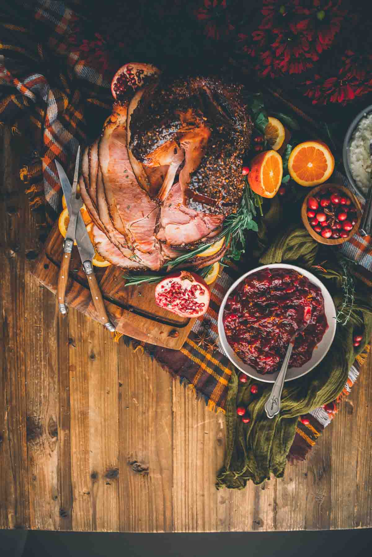 Overhead shot of holiday dinner table with sliced smoked ham and cranberry sauce, garnished with oranges, herbs and pomegranates.  