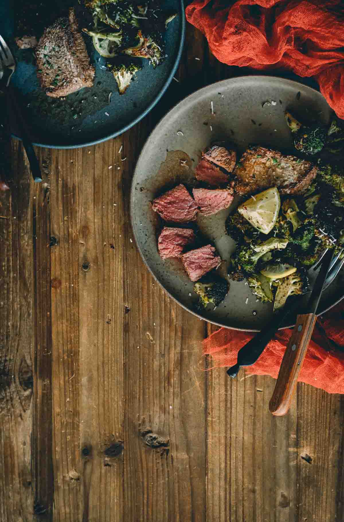 Overhead shot of 2 plates with venison steaks, one sliced, lemon wedge and roasted broccoli.