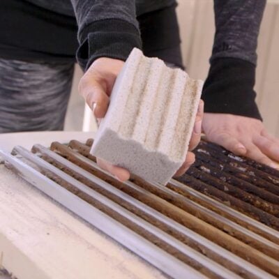 A person holds a textured cleaning block over rusty grill grates, showcasing an essential tool for grilling gifts enthusiasts who enjoy maintaining pristine cooking surfaces.