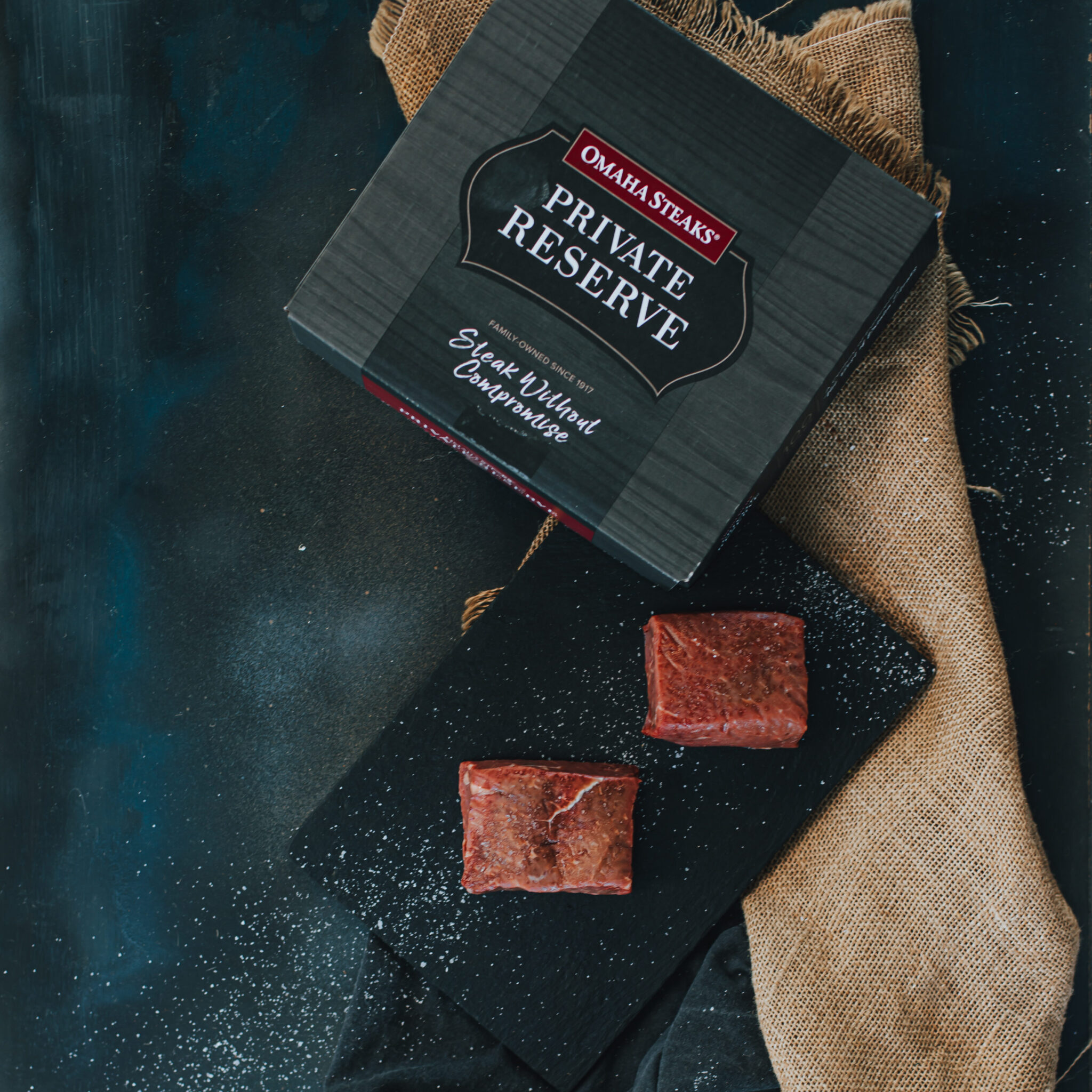 Overhead image of Omaha steaks box with two top sirloin filets on slate next to it to show packaging. 