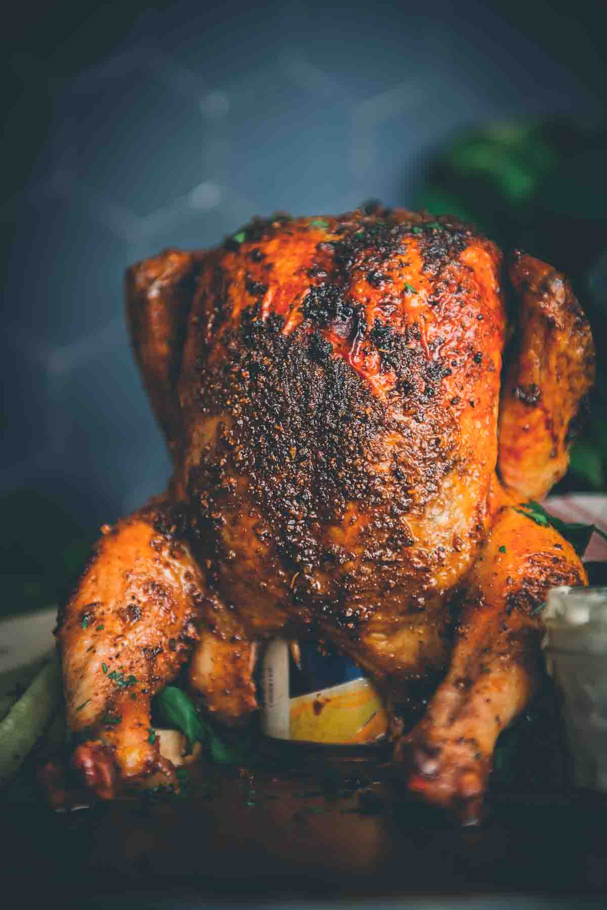 Smoked chicken with golden brown skin standing atop a beer can. 
