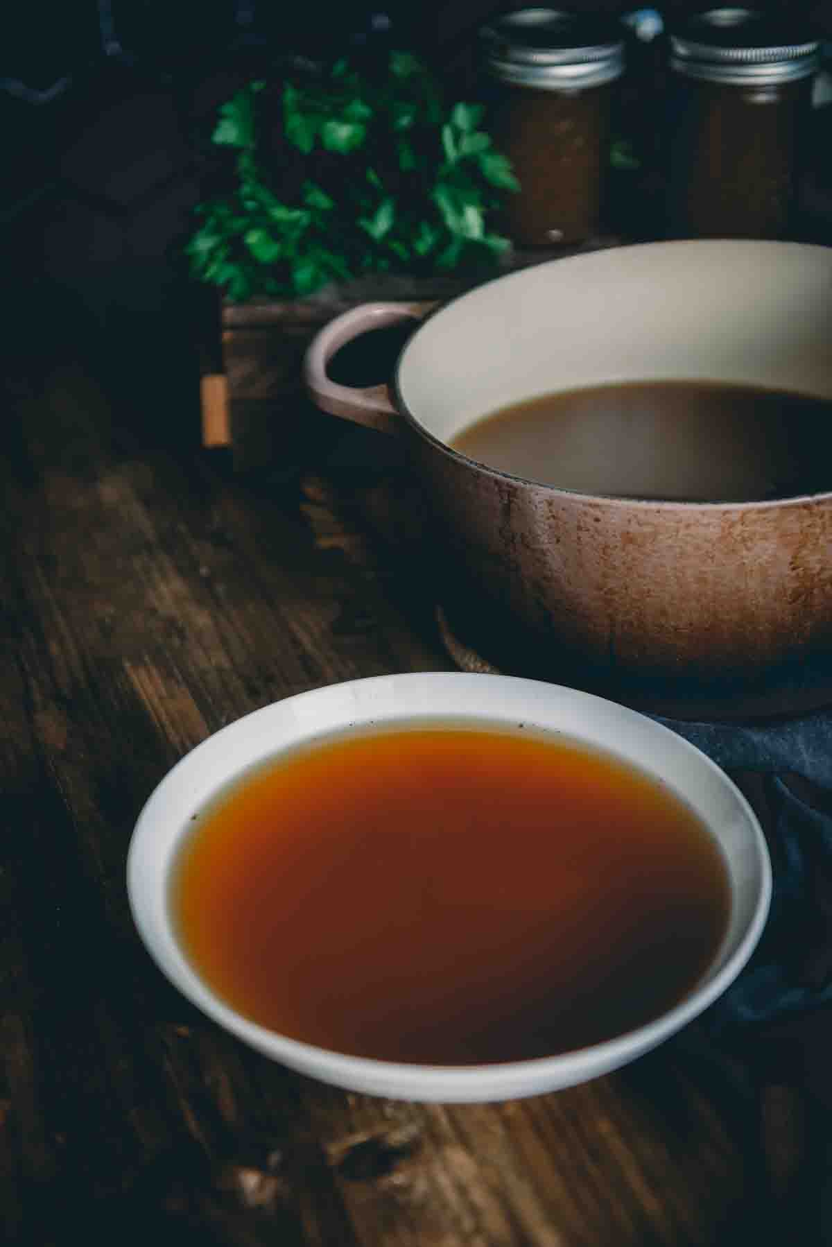 Close up of steaming bone broth in a white bowl on a wooden table with a dutch oven behind it. 