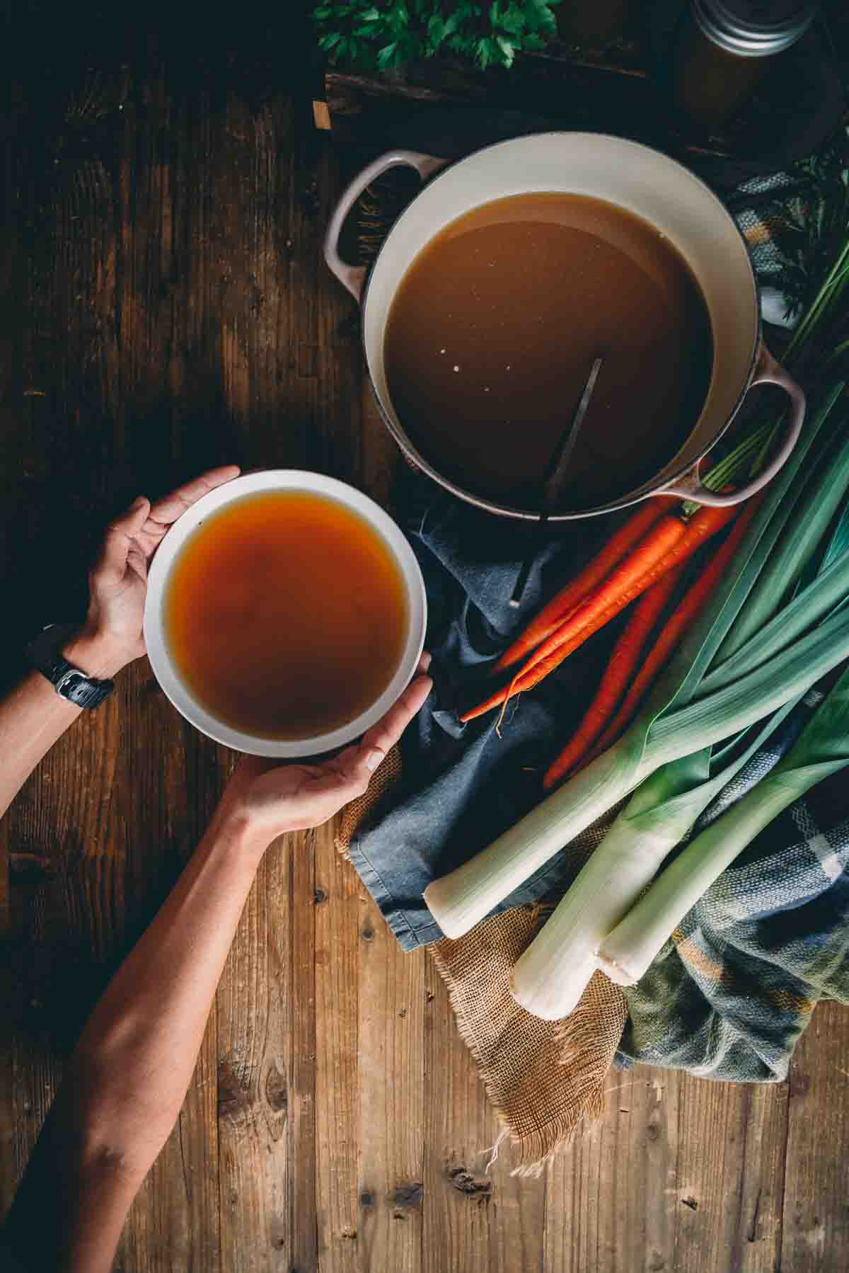 Hands holding bowl of bone broth over table of veggies. 