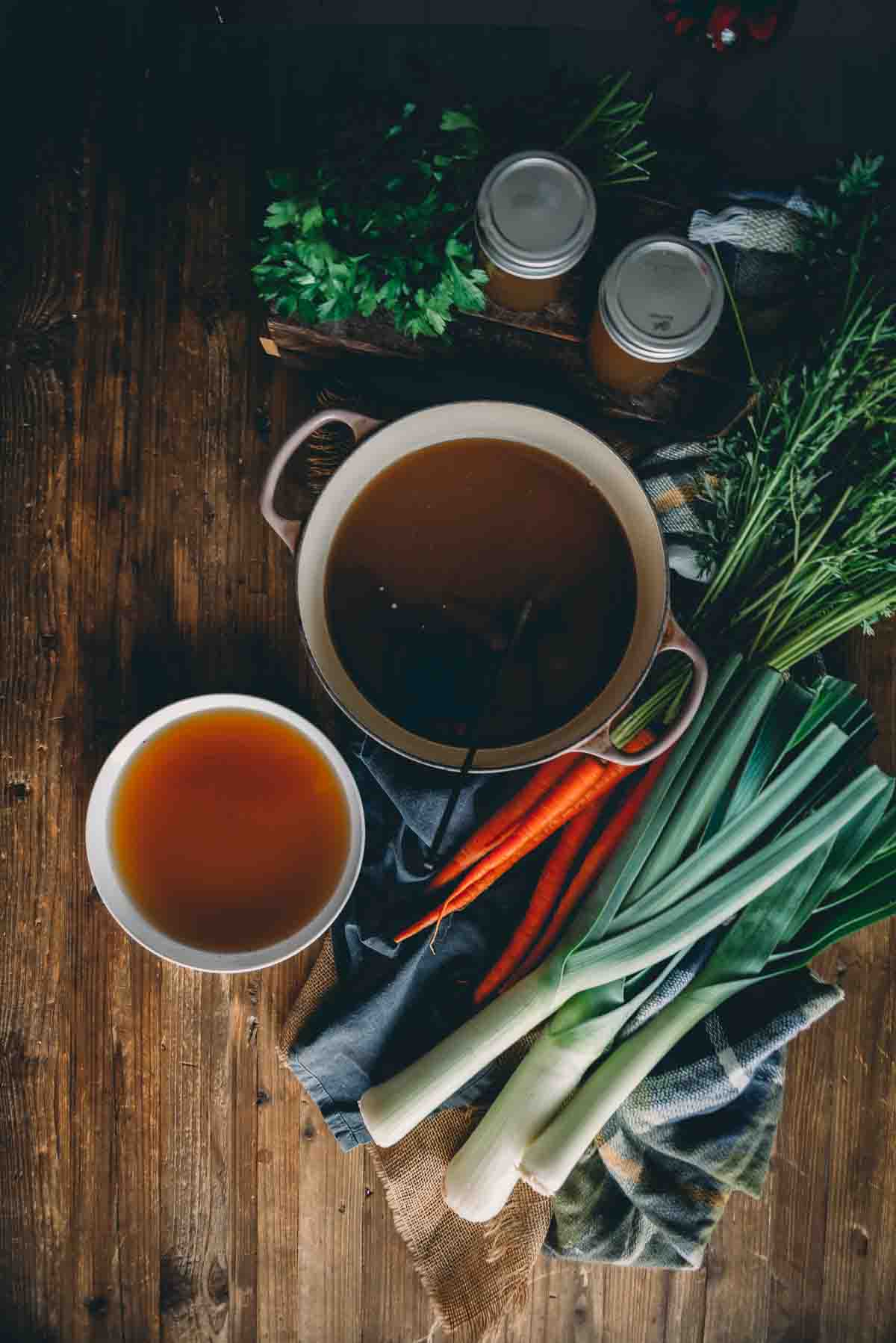 Overheads shot of dutch oven with bone broth and bowl filled with steaming broth on table with veggies. 