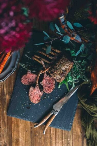 Rack of venison on a wooden board with carrots and herbs.