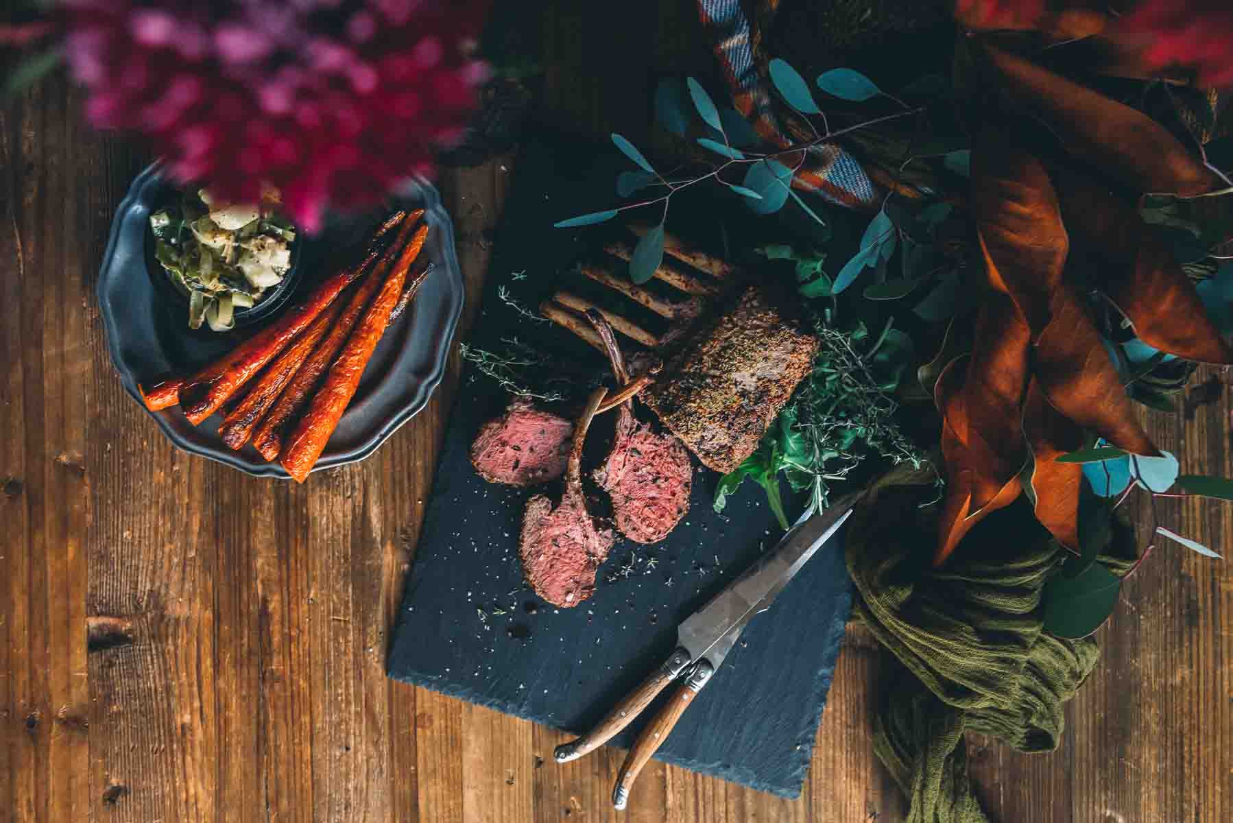 Overhead shot of rack of venison, with portions slices on a slate board with roasted carrots and leeks nearby on a wooden table. 