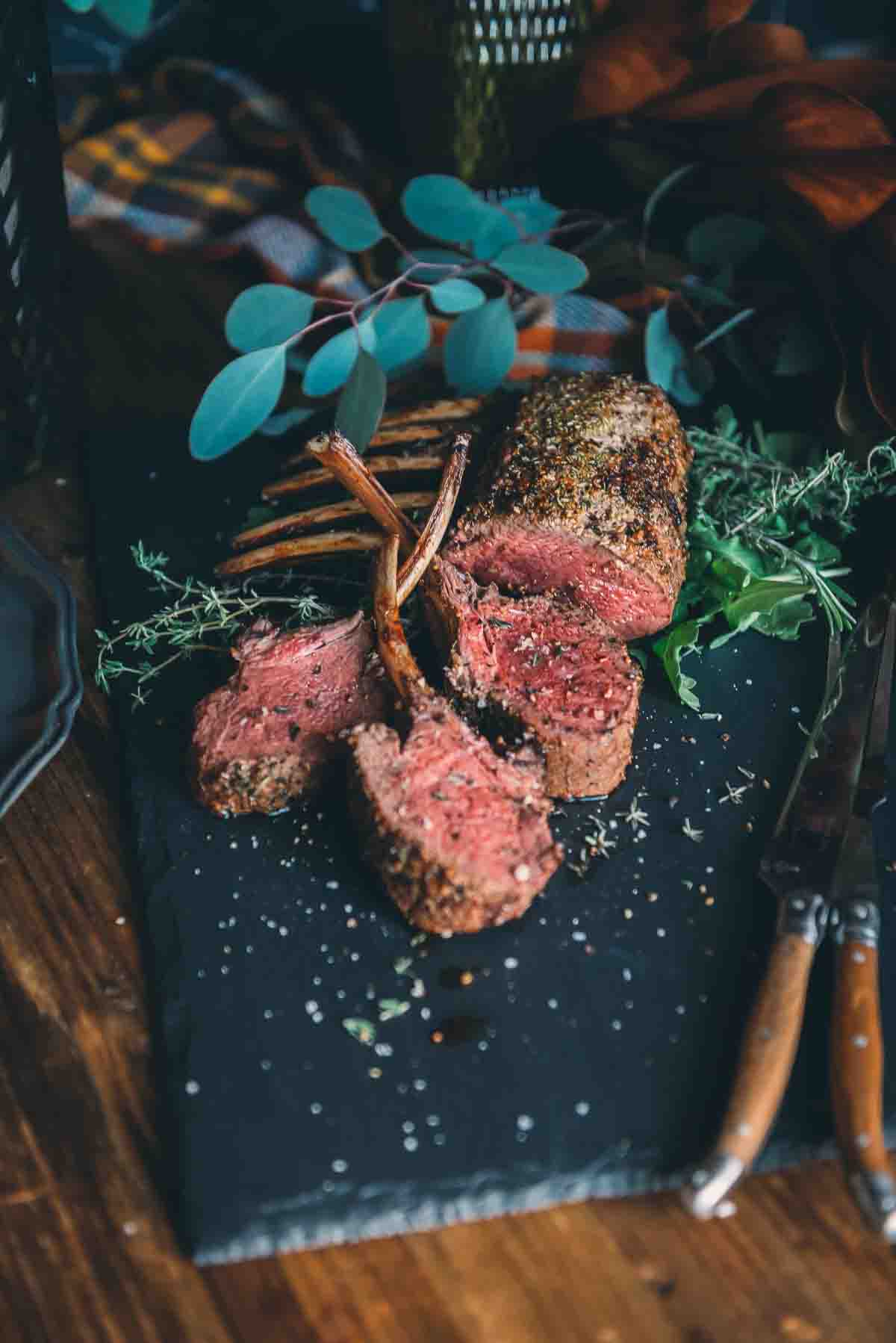 Showing a rack of venison with three slices carved on a slake board with carving fork and knife and herbs. 